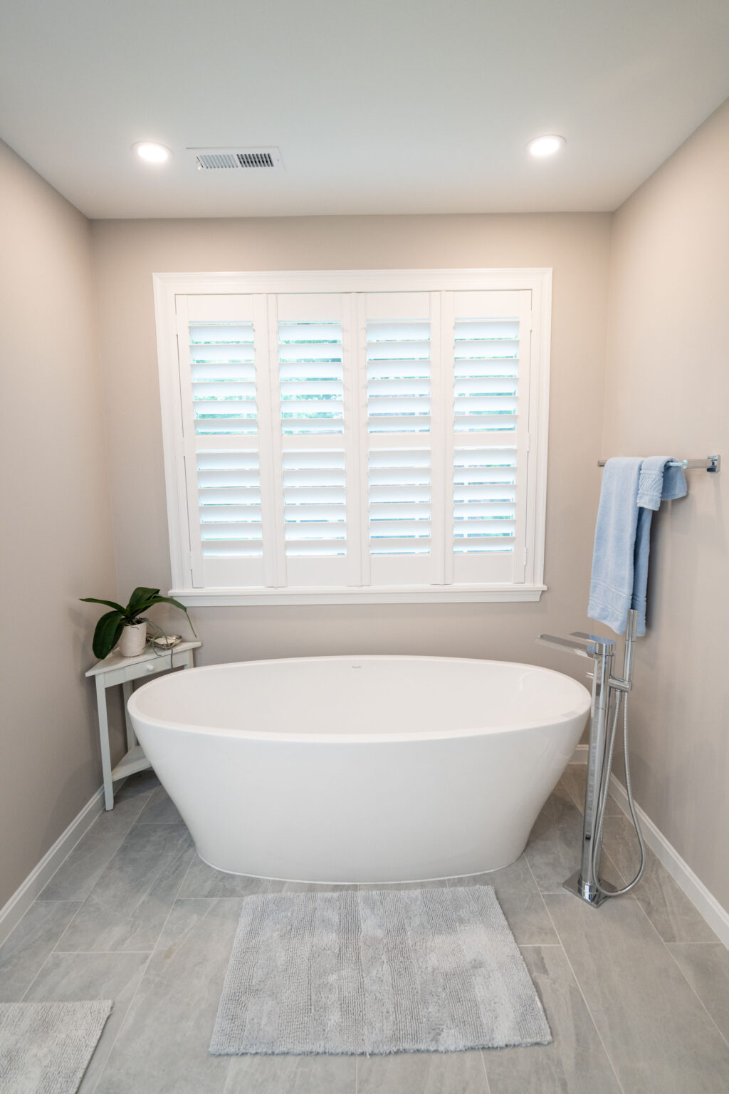 A modern bathroom with a freestanding white tub, a white side table with a plant, and a towel rack with a blue towel. White shutters cover the window above the tub. The floor is tiled in light gray.