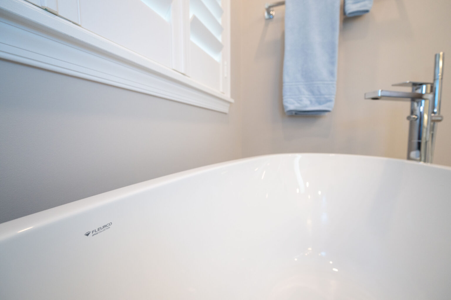 Close-up view of a white bathtub with a chrome faucet next to a towel rack holding a light blue towel, against a beige wall and white window shutters.