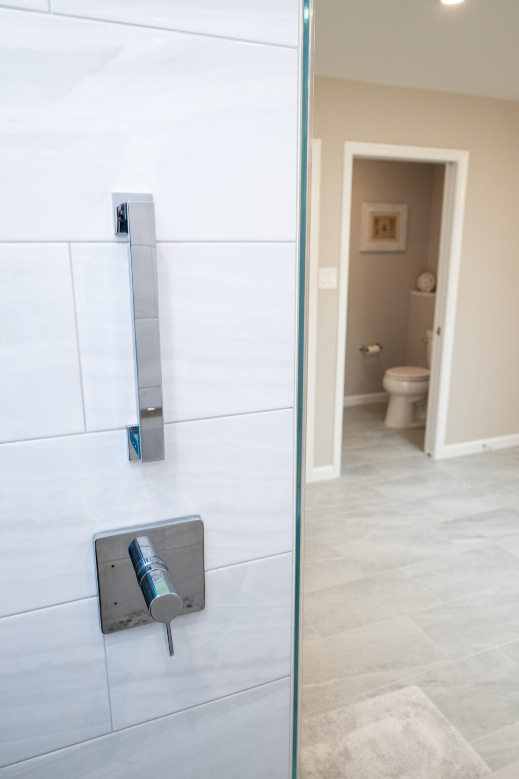 Bathroom with tiled walls featuring a modern chrome shower handle in the foreground. An open doorway in the background reveals a toilet and beige wall with a small wall-mounted clock.