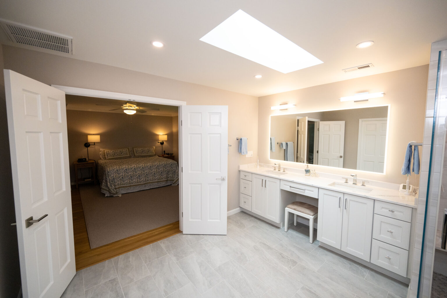 A bathroom with a large mirror and double sink vanity. The doorway leads to a bedroom with a bed, nightstands, and lamps. The bathroom has tile flooring and the bedroom has carpeted flooring.