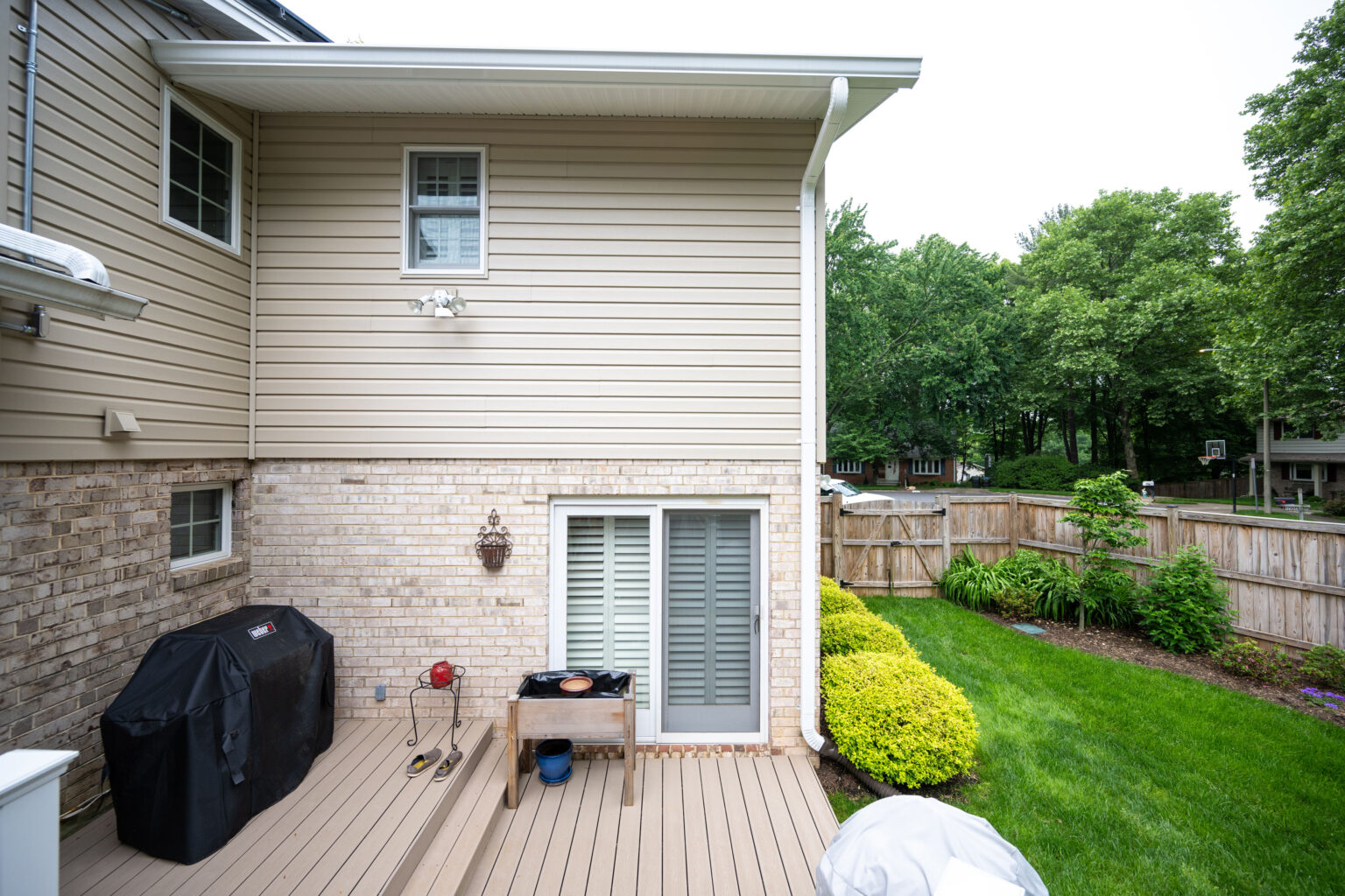 Backyard view featuring a wooden deck with a grill, a small table, and a fenced-in yard with green grass and bushes along the perimeter. The house has light-colored siding and brick with a patio door.