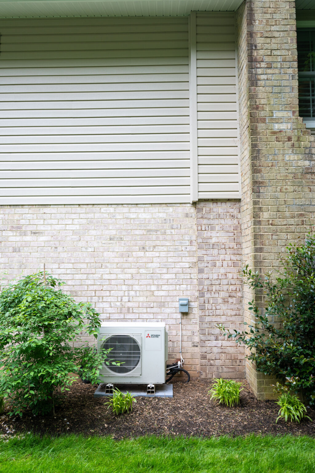 A residential air conditioning unit is mounted on a concrete pad outside a house, surrounded by landscaped bushes and plants, with brick and siding on the house wall.