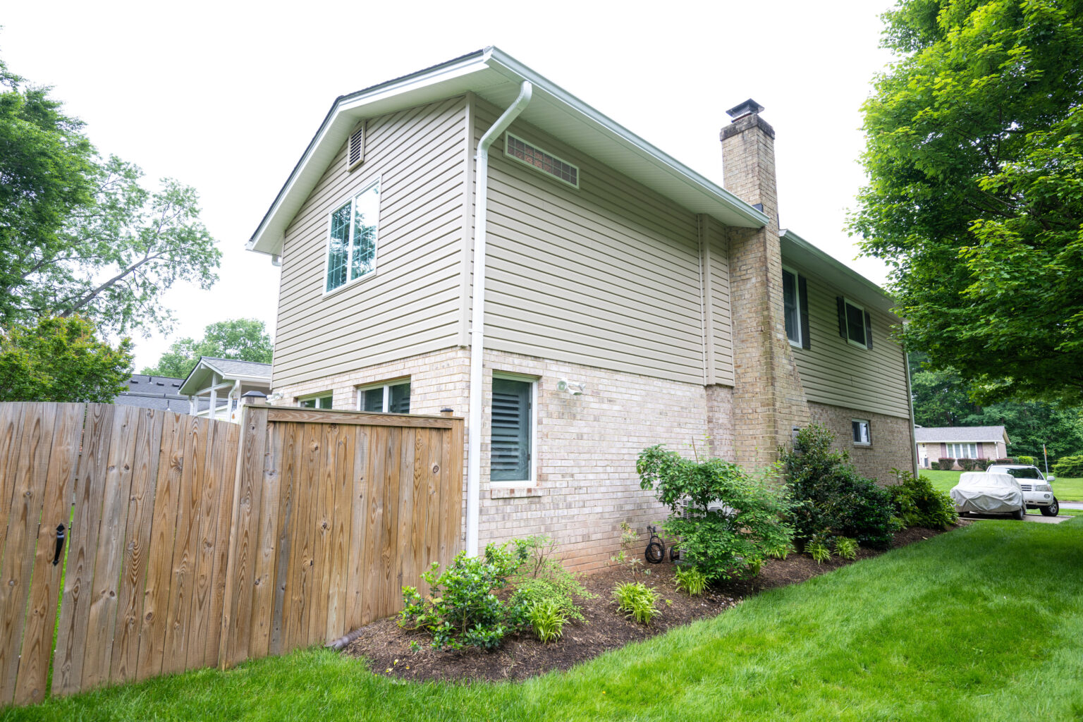 A two-story house with beige siding and a brick lower section, adjacent to a wooden fence and landscaped garden, surrounded by green lawn and trees.