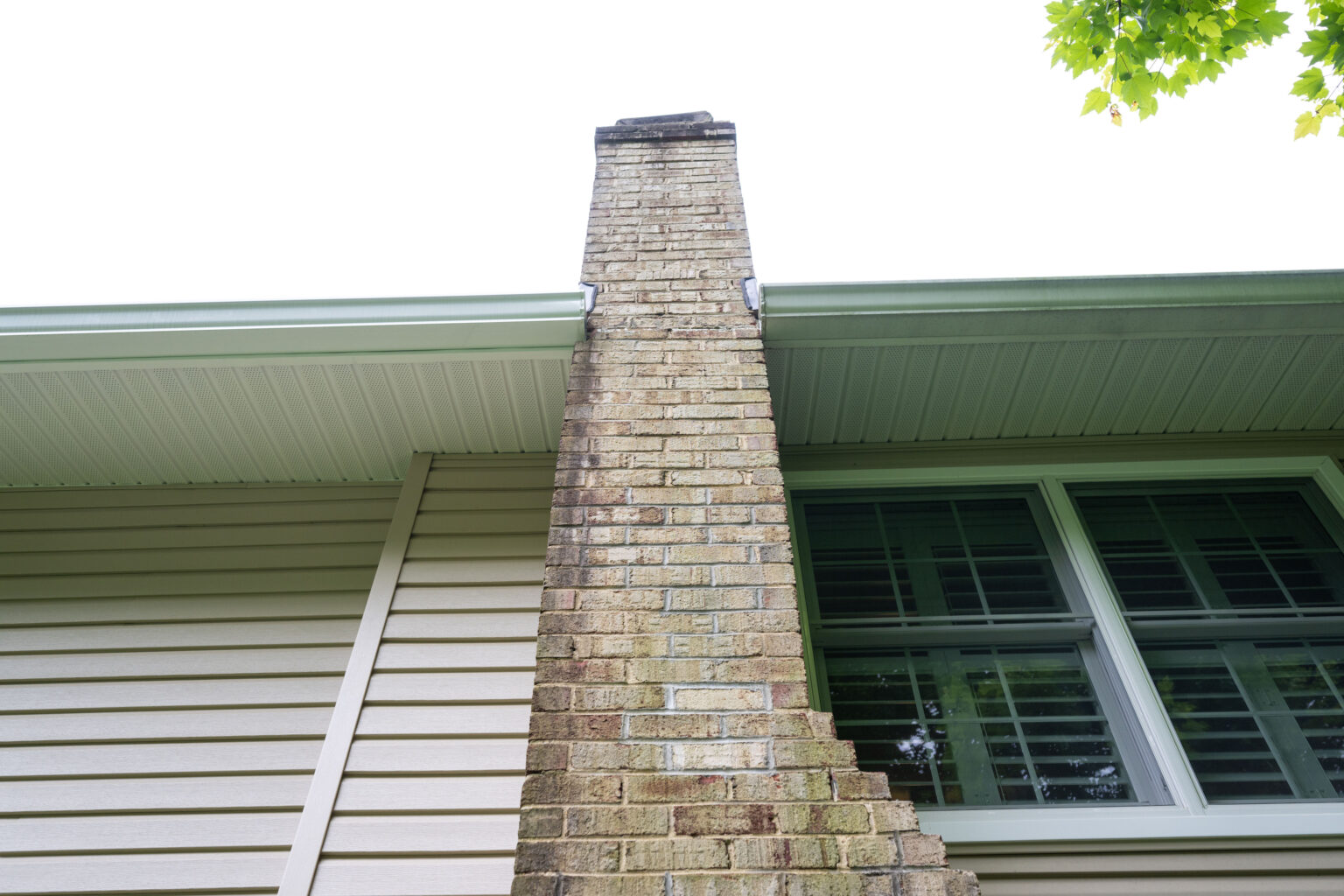 View from below of a brick chimney extending from the roofline of a house with beige siding and large windows.
