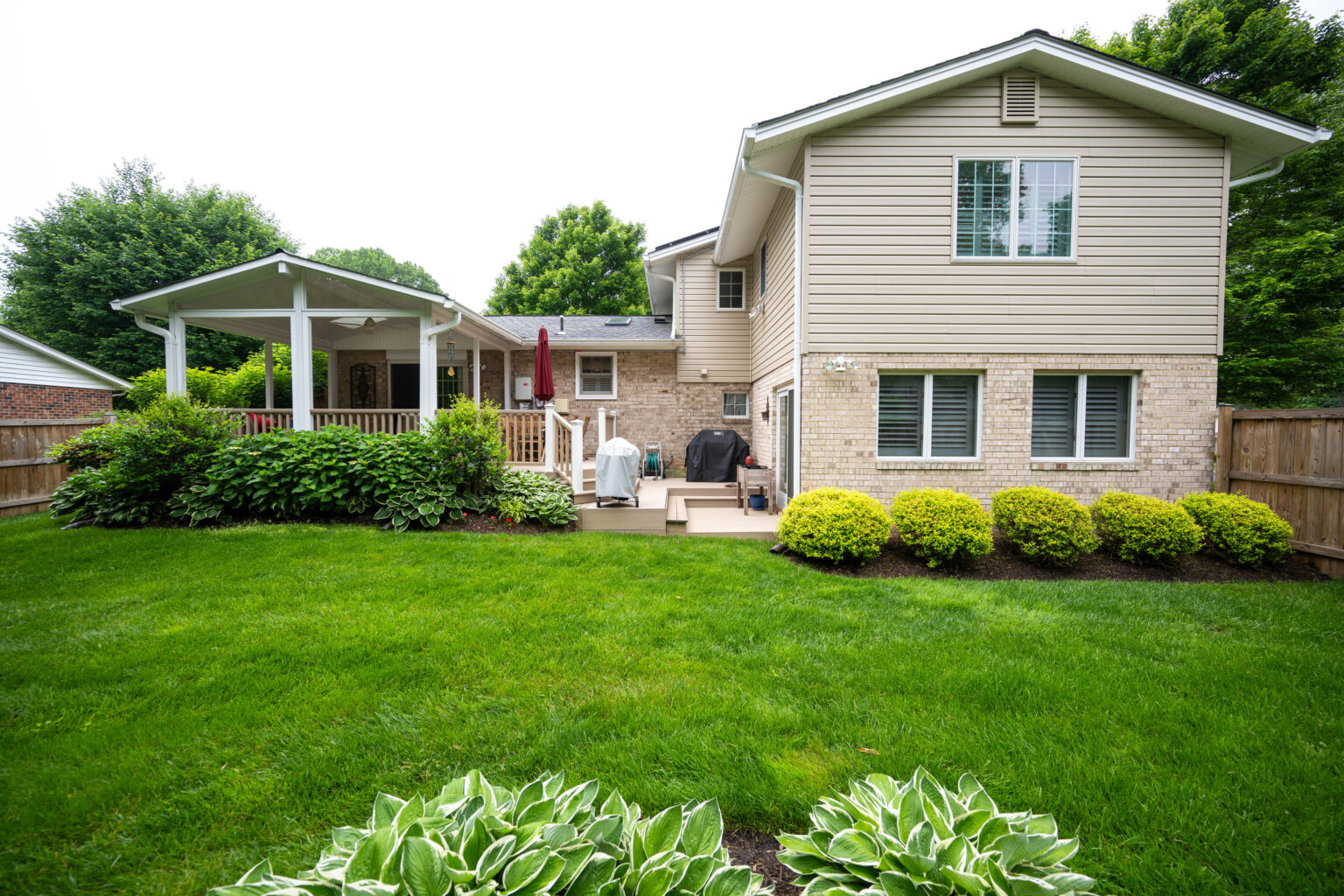 A backyard with a green lawn, a wooden deck with a barbecue grill, a covered patio area, and a beige house with multiple windows. Bushes and plants line the edge of the deck and fence.