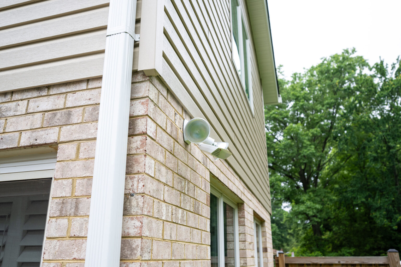 A floodlight is mounted on the brick wall of a beige, two-story house. A white downspout runs down the corner of the house, and trees are visible in the background.