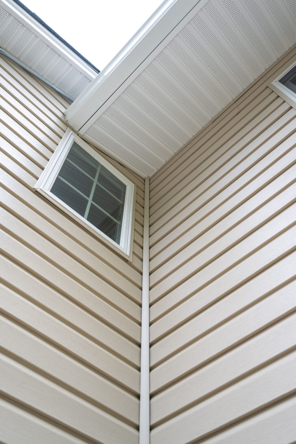 Close-up view of the exterior of a beige house corner showing horizontal siding, a window with white trim, and a gutter running vertically between rooflines.