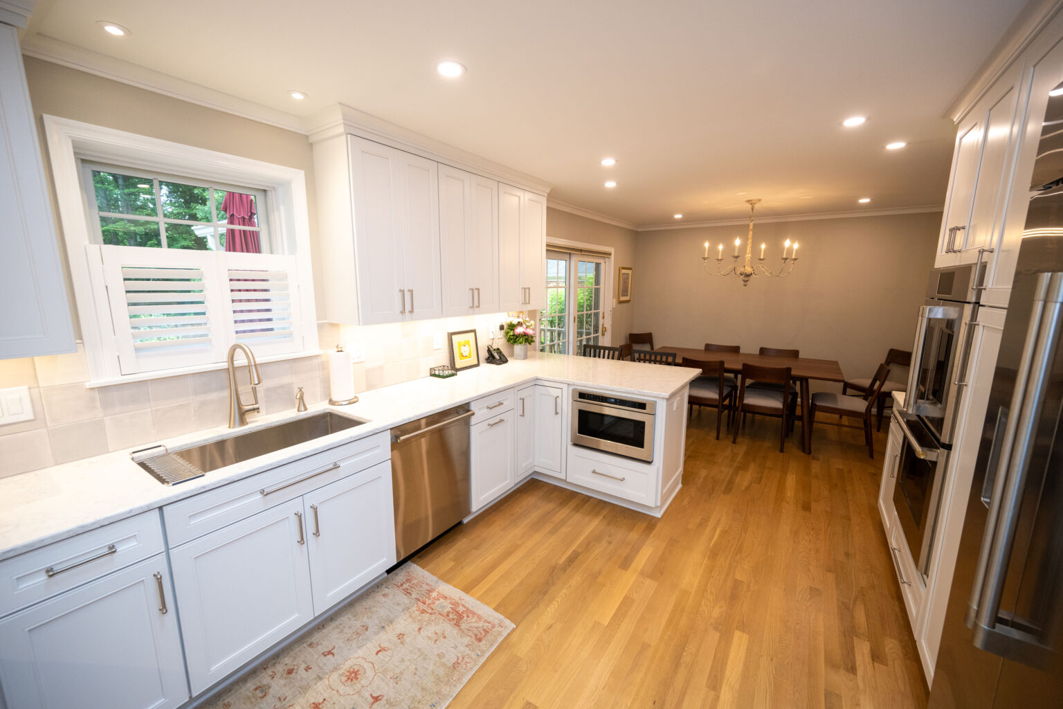 Image by Lesnick Photo A modern kitchen with white cabinets, stainless steel appliances, and hardwood floors opens to a dining area with a wooden table, chairs, and a chandelier.