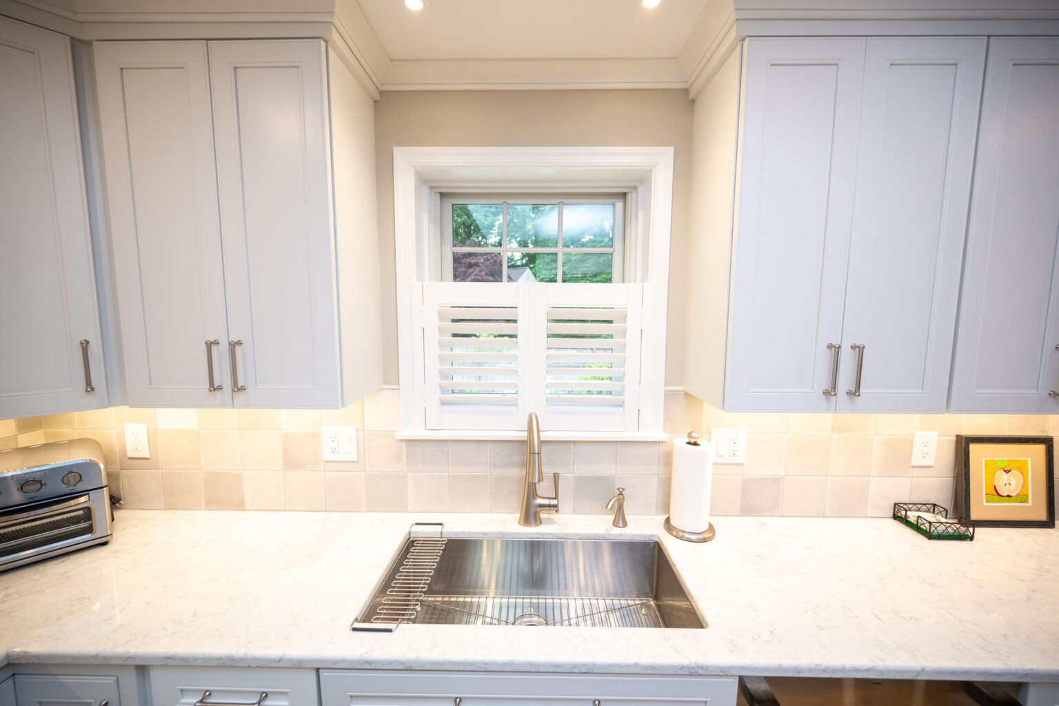 Image by Lesnick Photo A modern kitchen sink area with white marble countertops, gray cabinets, and a window above the sink, partially covered by white shutters. Includes a toaster, paper towel holder, and framed art.