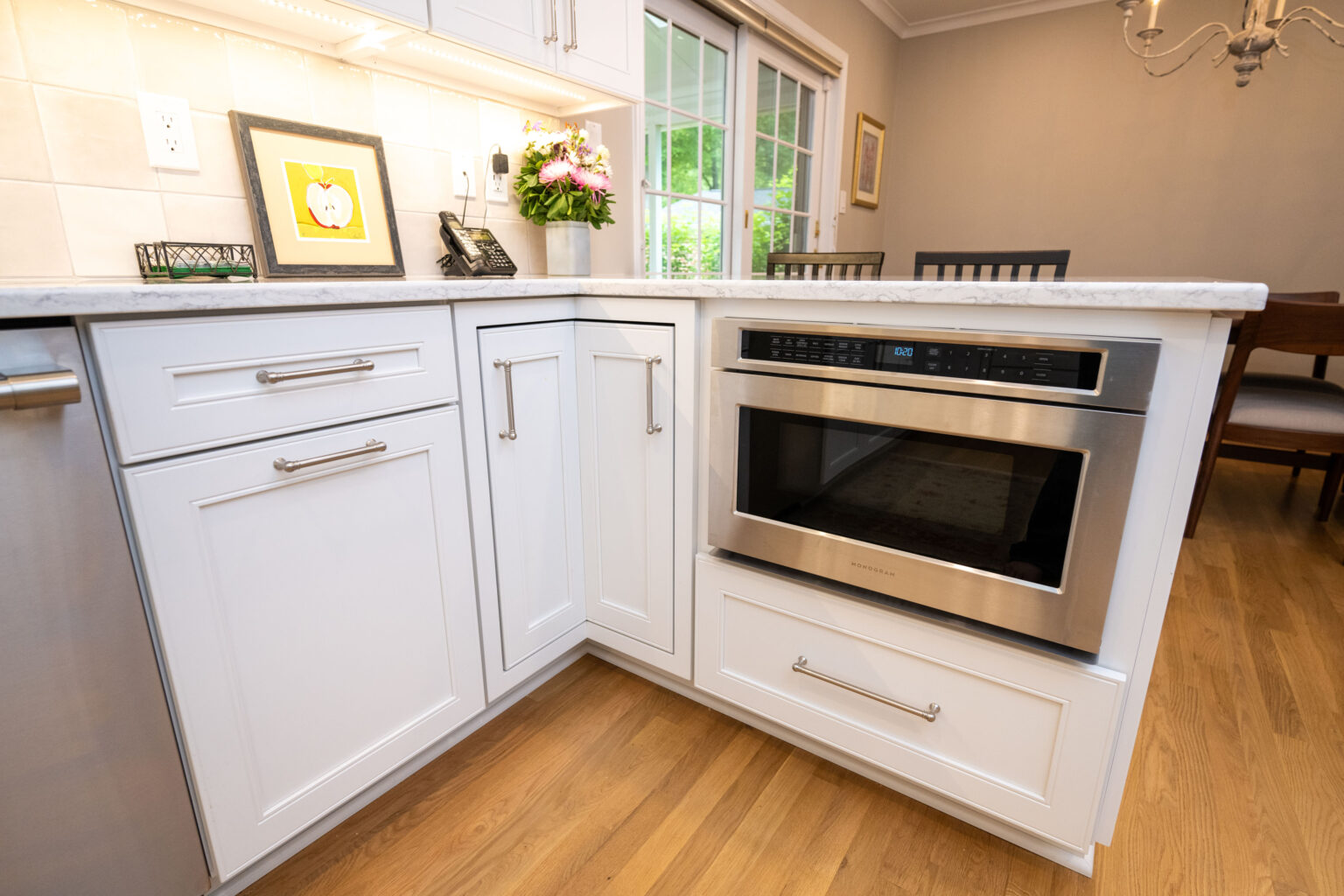 Image by Lesnick Photo A modern kitchen corner with white cabinets, stainless steel appliances, a small flower arrangement, and a framed picture on the countertop. Large windows in the background provide natural light.
