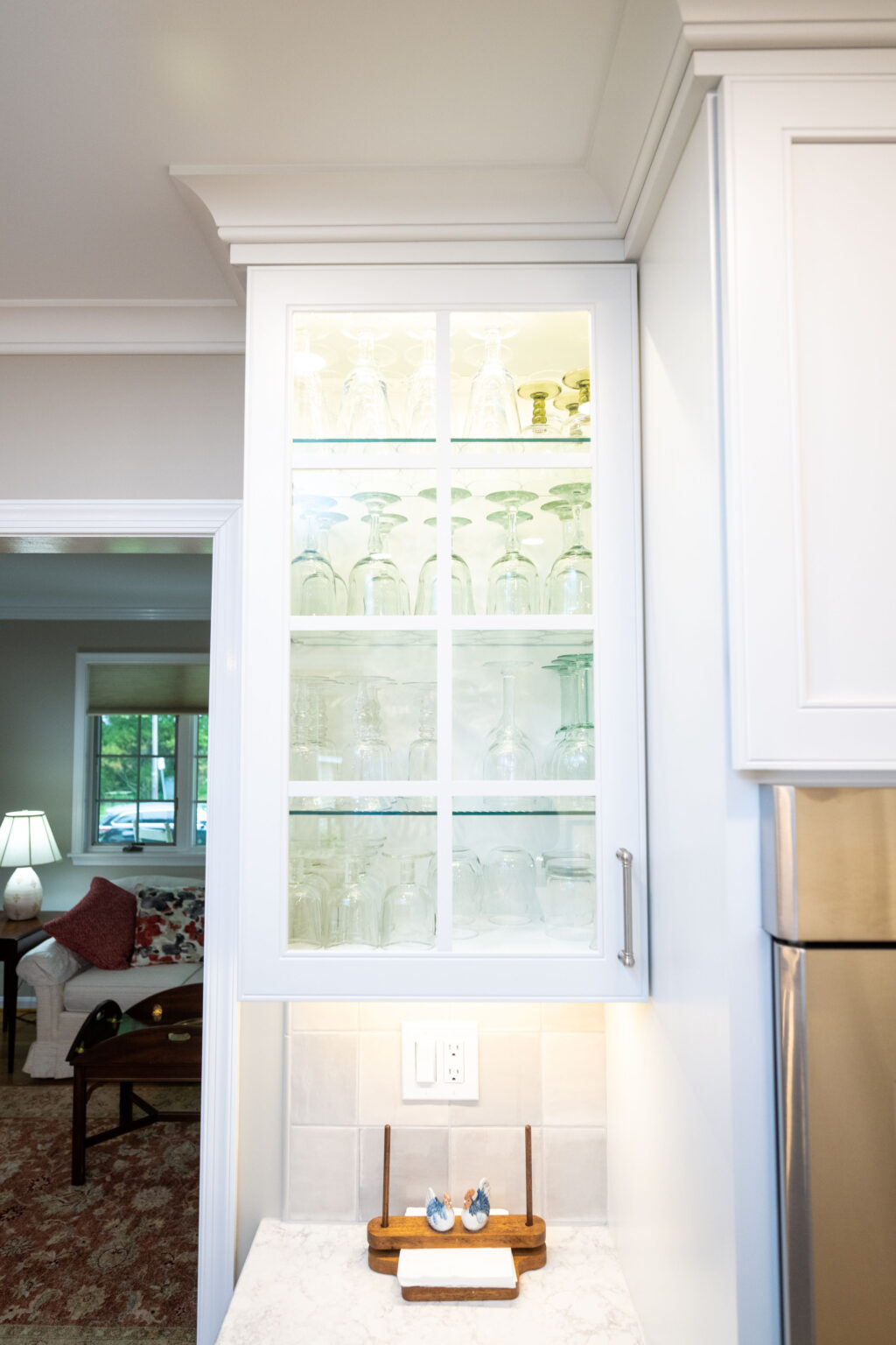 Image by Lesnick Photo A kitchen cabinet with glass-paneled doors displays neatly arranged glassware. The countertop below has a small tray with salt and pepper shakers and an electrical outlet above it.