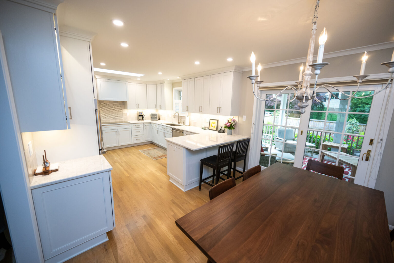 Image by Lesnick Photo A modern kitchen and dining area with white cabinets, a wooden dining table, a chandelier, and an island. Large windows provide natural light and a view of an outdoor seating area.