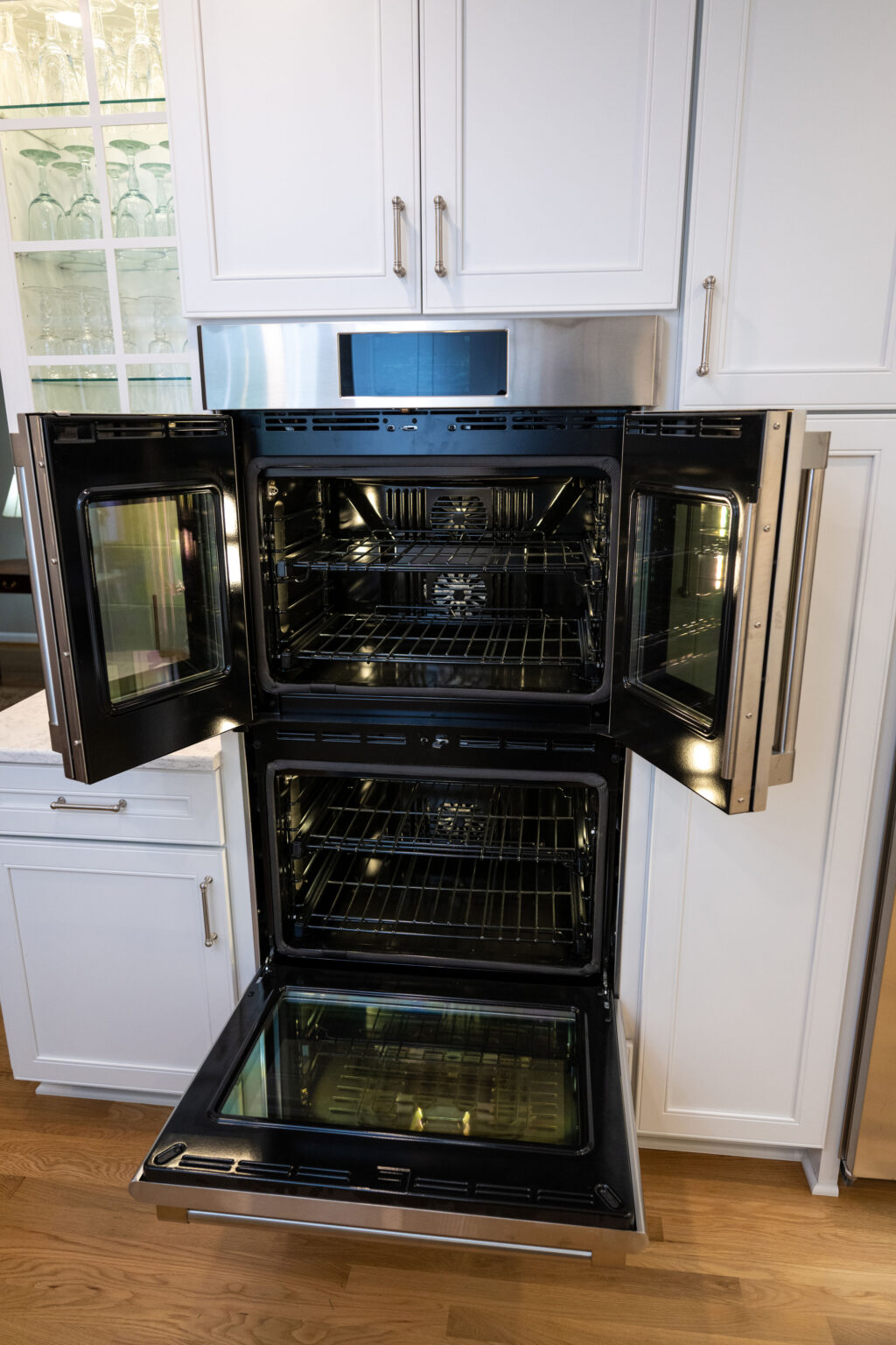 Image by Lesnick Photo A modern kitchen features a built-in double oven with both doors open, revealing clean, empty oven interiors. White cabinetry and wooden flooring surround the stainless steel appliance.