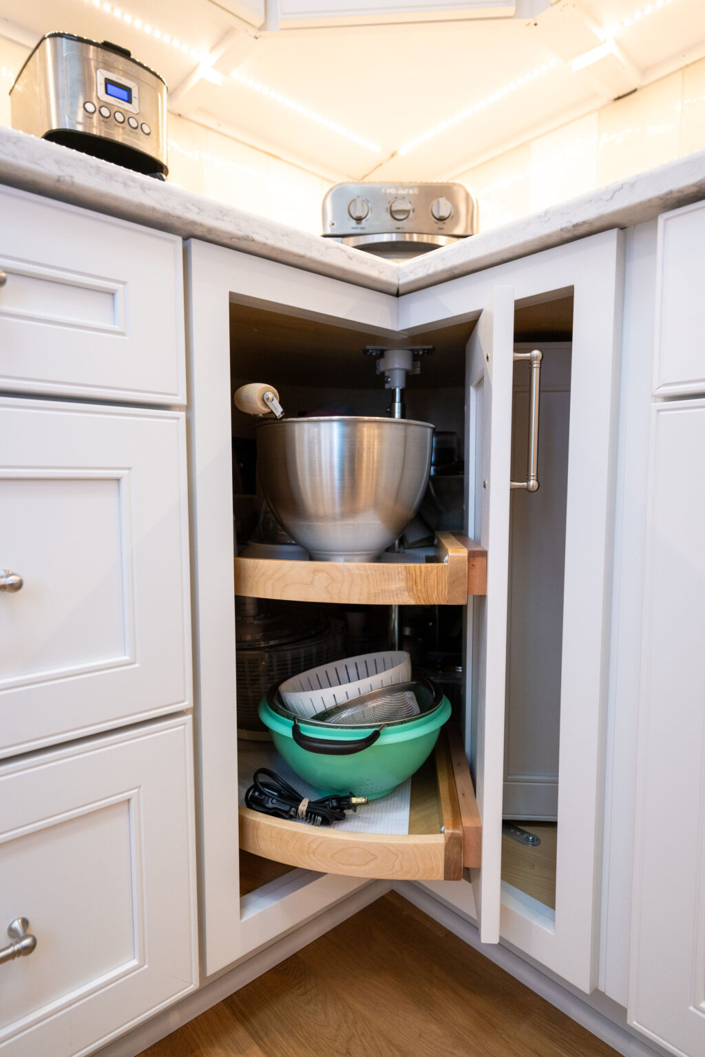 Image by Lesnick Photo Corner kitchen cabinet with two pull-out wooden shelves. The top shelf holds a large mixing bowl, and the lower shelf contains a green colander and other kitchen items. White cabinets and a toaster on the counter.