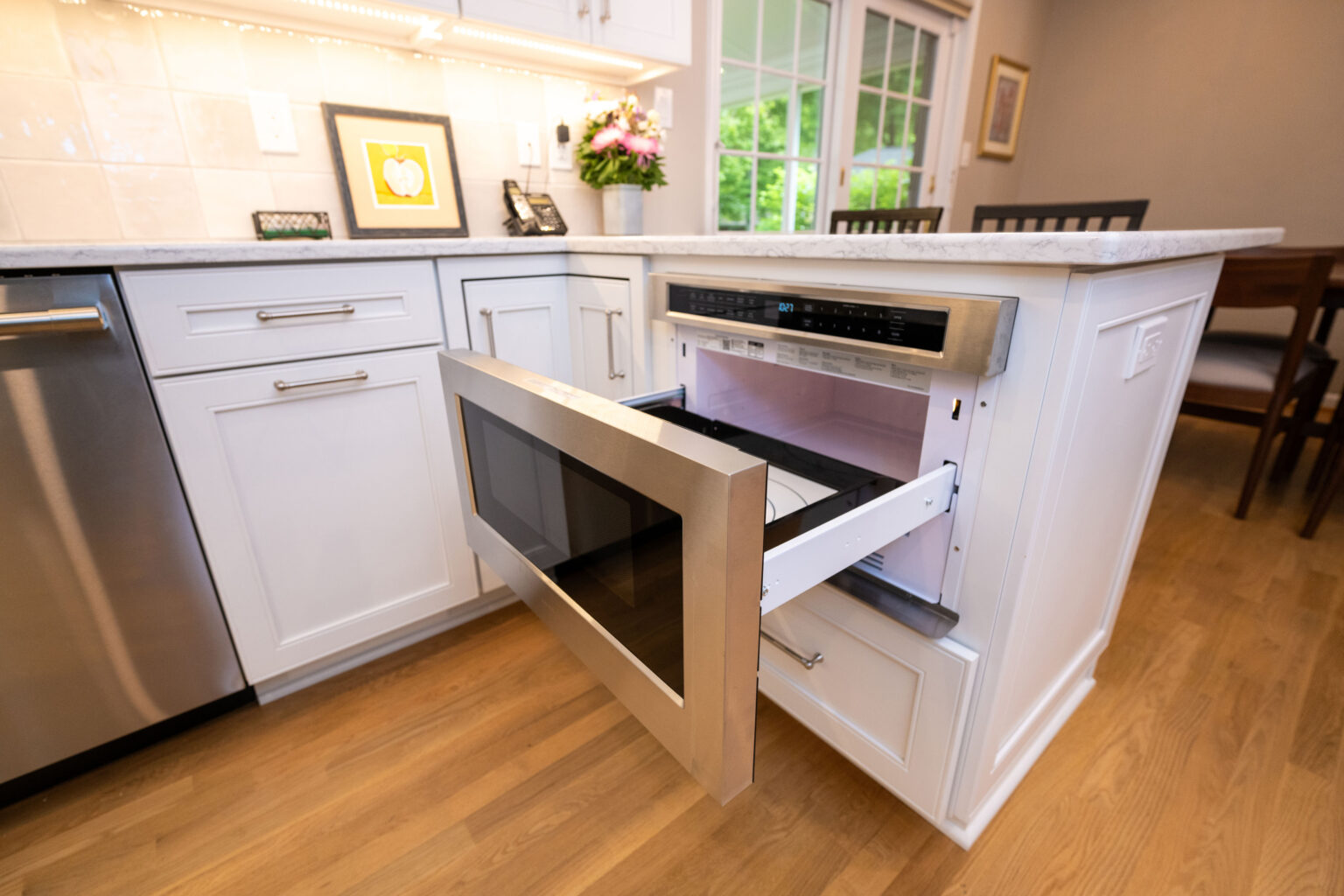 Image by Lesnick Photo A kitchen with white cabinets, a stainless steel dishwasher, and an open drawer-style microwave installed beneath the countertop. There are flowers and framed artwork on the counter.