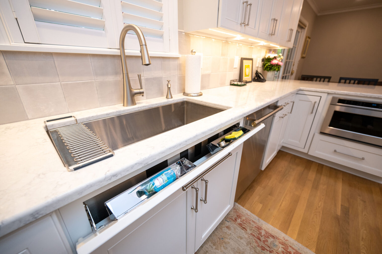 Image by Lesnick Photo A modern kitchen with white cabinets, stainless steel appliances, and a marble countertop. The sink has a drying rack, and one drawer is partially open, revealing utensils and cleaning supplies.