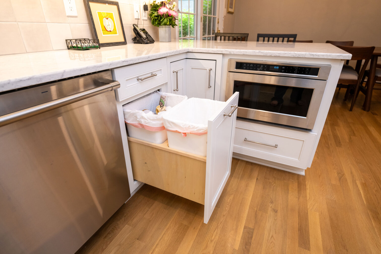Image by Lesnick Photo A modern kitchen corner featuring stainless steel appliances and white cabinets with an open pull-out trash and recycling bin drawer.