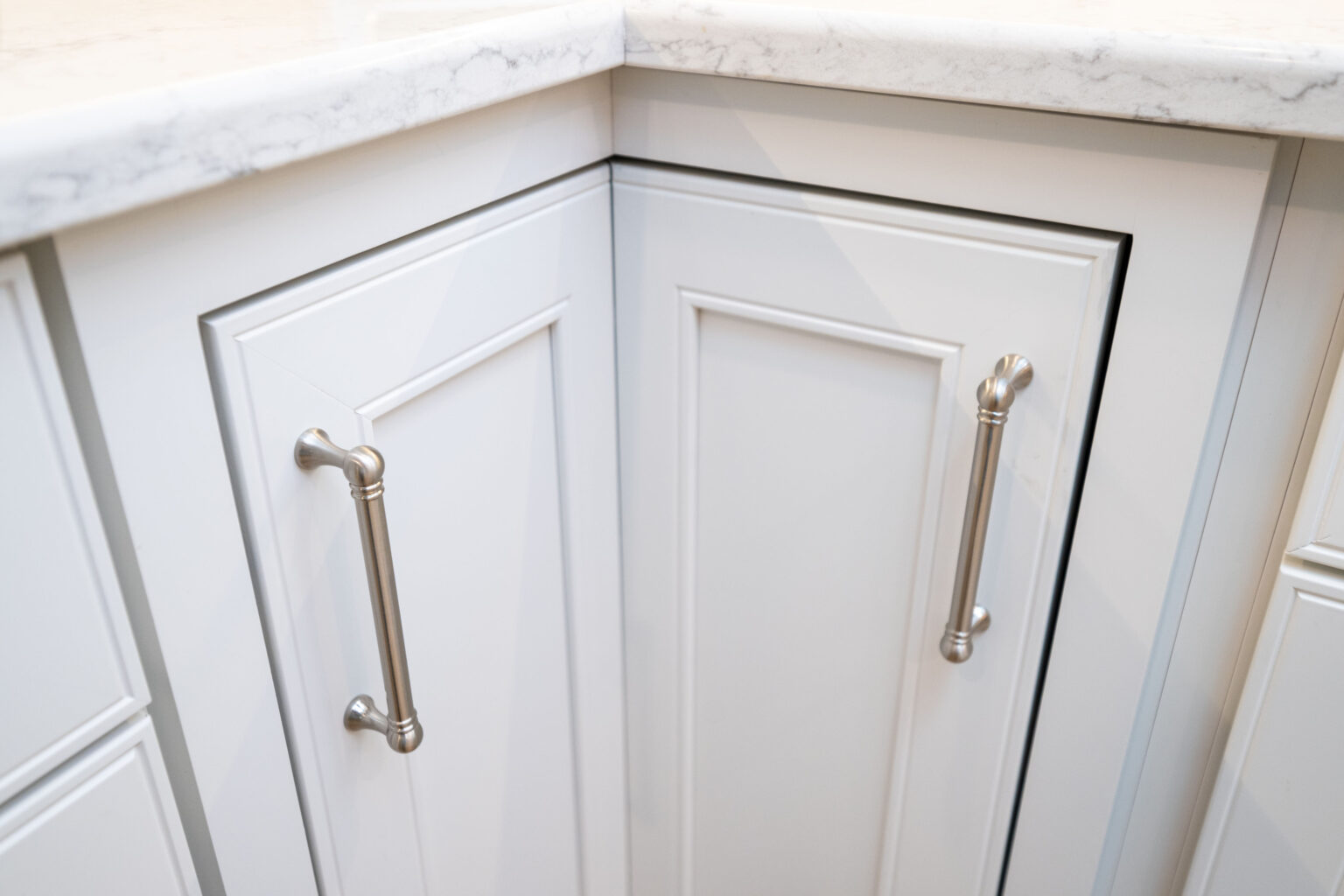 Image by Lesnick Photo Close-up of two white kitchen cabinet doors at a corner with silver handles and a marble countertop above them.