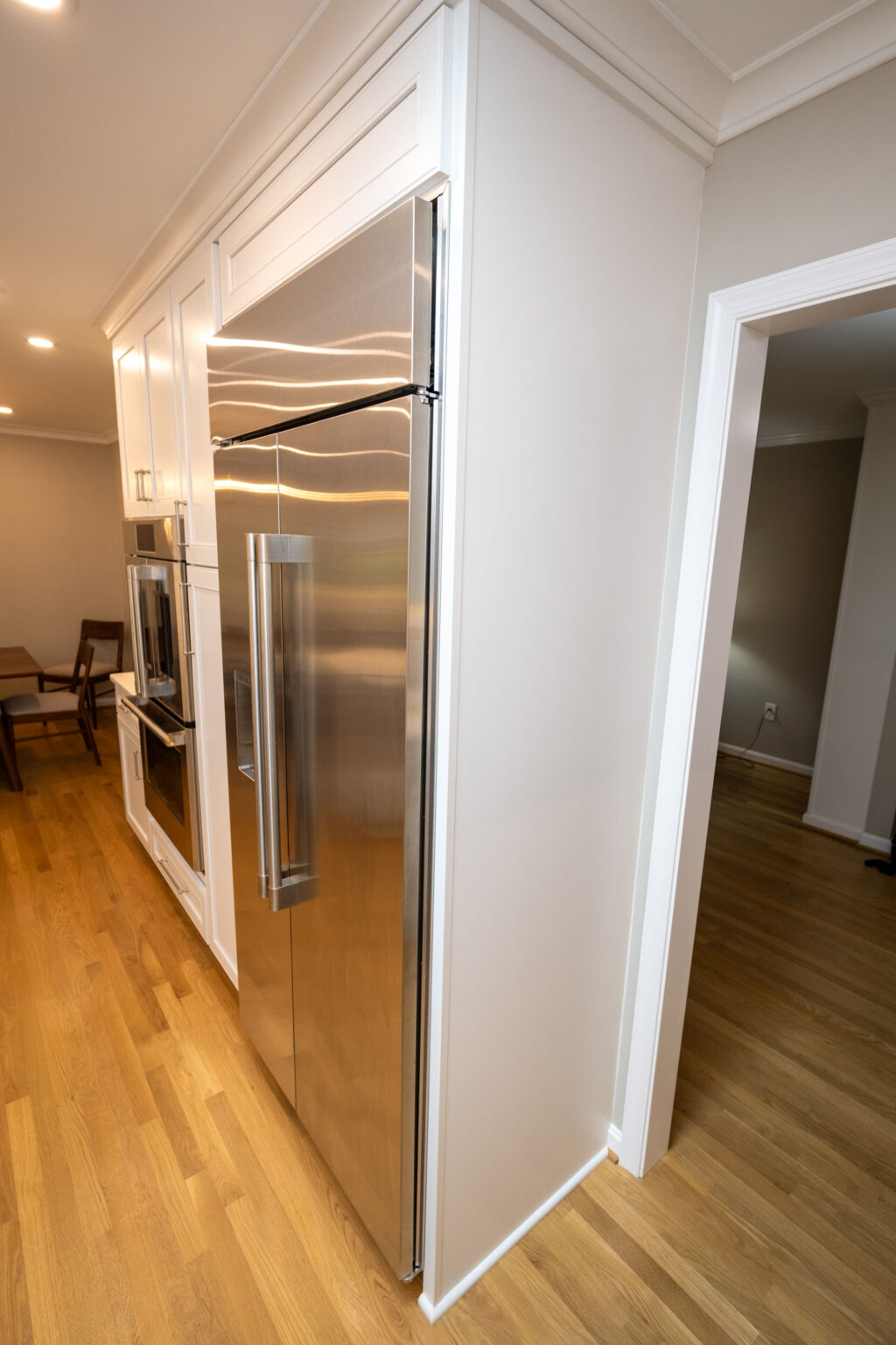 Image by Lesnick Photo A modern stainless steel refrigerator is installed in a well-lit kitchen with wooden floors, white cabinetry, an oven, and a dining table visible in the background.