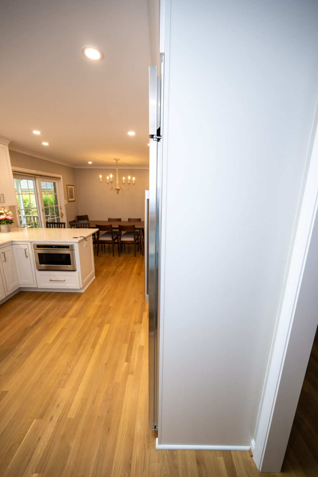 Image by Lesnick Photo A modern kitchen with wooden flooring, white cabinets, a built-in microwave, and a dining area with a chandelier in the background.