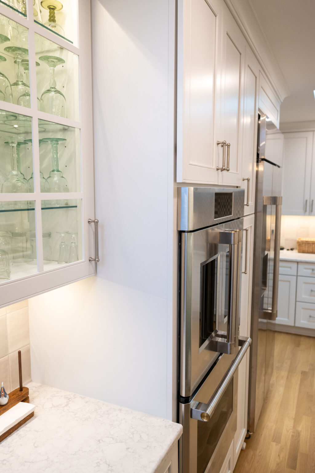 Image by Lesnick Photo A modern kitchen with white cabinetry, glass-front cabinets displaying glassware, a stainless steel double oven, and a marble countertop.
.