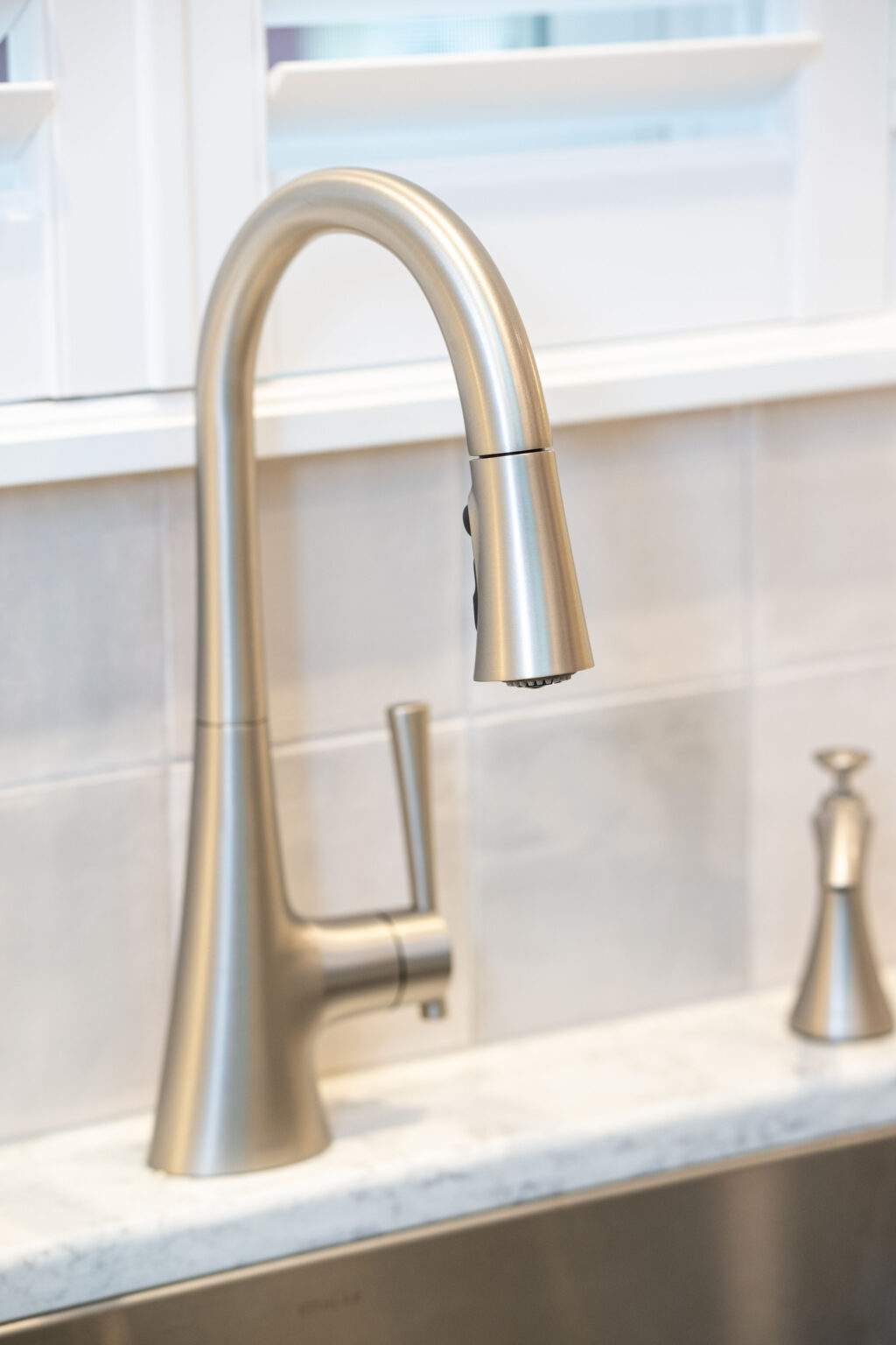 Image by Lesnick Photo A modern stainless steel kitchen faucet with a curved spout and a single lever handle, installed on a light-colored countertop against a tiled backsplash.