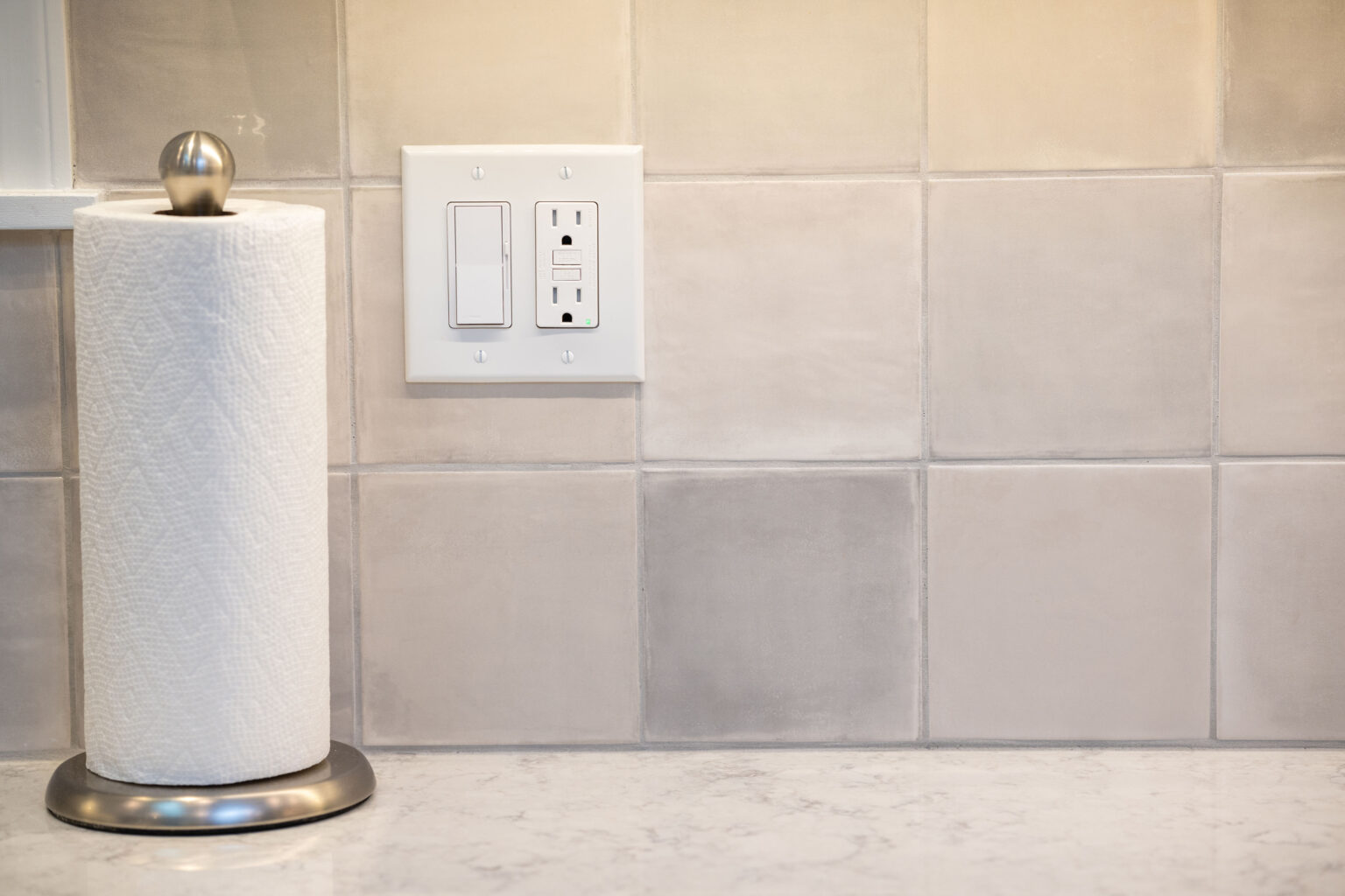 Image by Lesnick Photo A roll of paper towels stands on a holder next to a wall outlet with two sockets and a switch, mounted on a light-colored tiled backsplash.