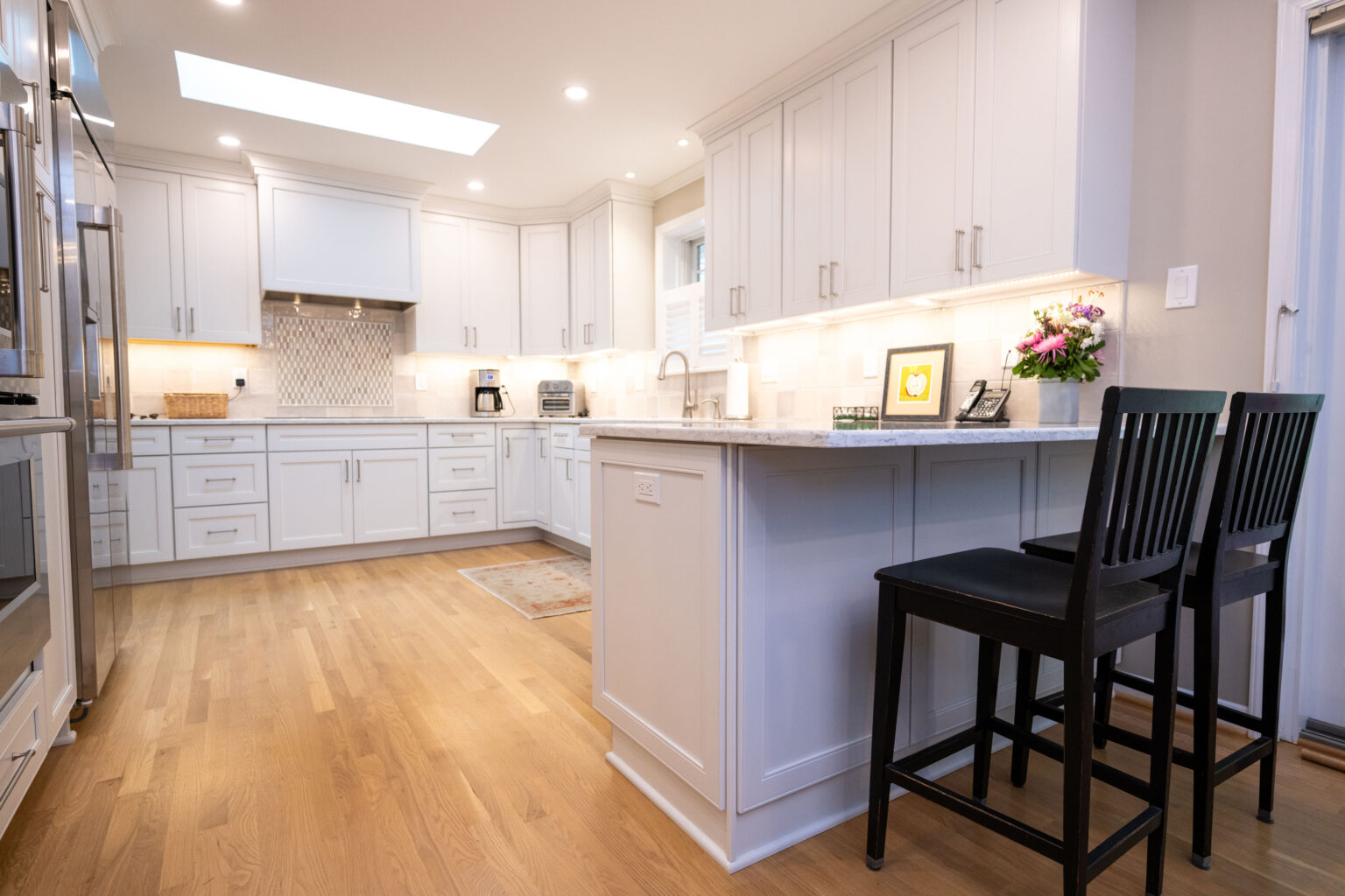 Image by Lesnick Photo A modern kitchen with white cabinets, a center island with two black chairs, stainless steel appliances, and flowers on the counter. The room has hardwood floors and a skylight.