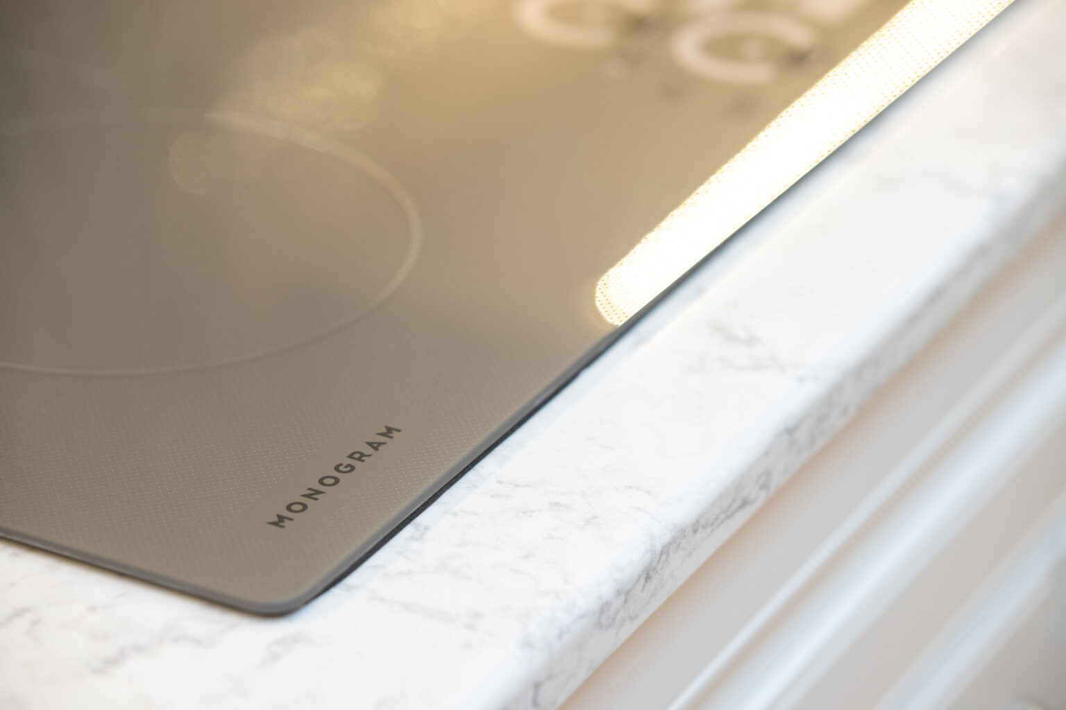 Image by Lesnick Photo A close-up of a kitchen countertop showing a sleek, glass-ceramic induction cooktop with the word "MONOGRAM" visible at the edge. Countertop appears to be made of white marble or similar material.