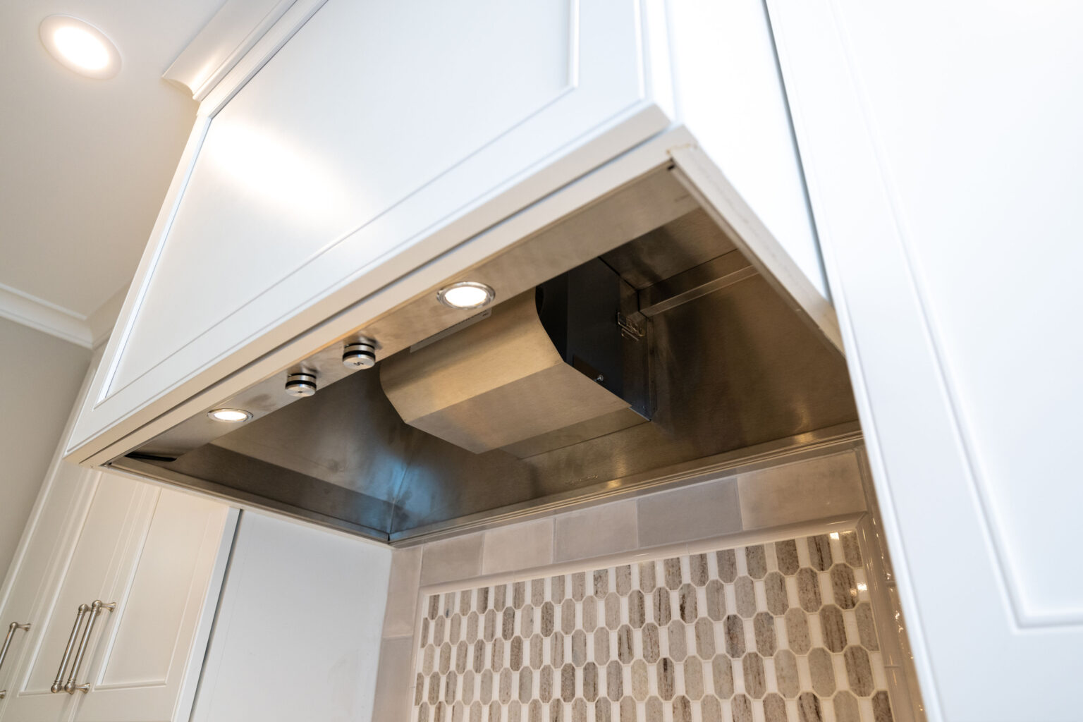 Image by Lesnick Photo Close-up view of a modern kitchen range hood with stainless steel finish and built-in lights, installed above a tiled backsplash.