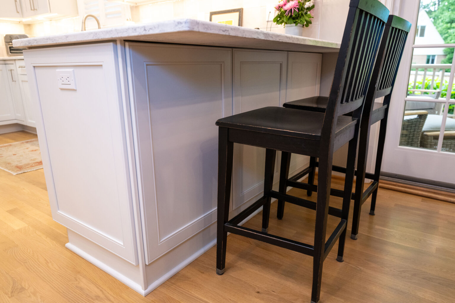 Image by Lesnick Photo A kitchen island with a white marble countertop and two black wooden chairs on a light wood floor, located near a window.