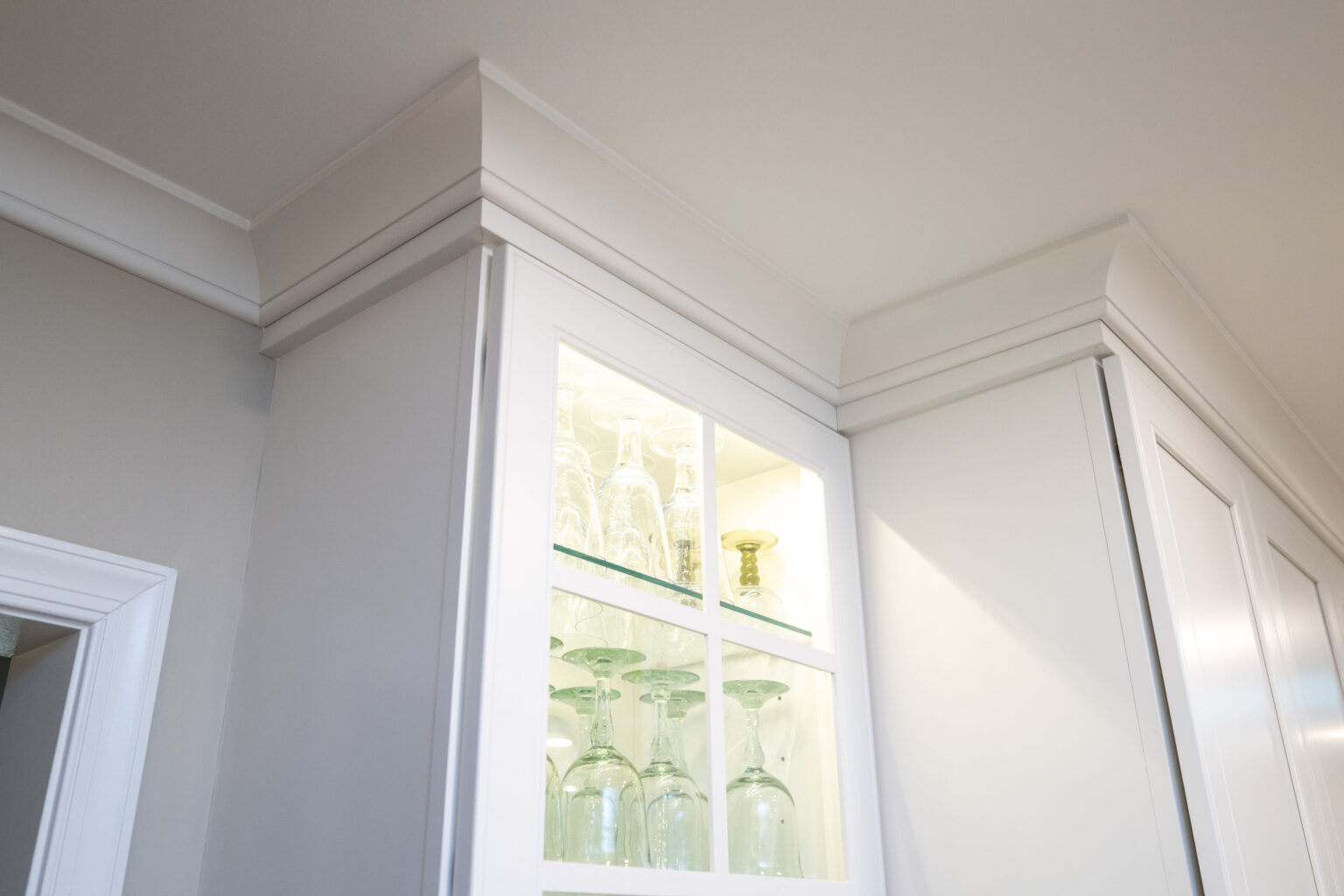 Image by Lesnick Photo A close-up of a white kitchen cabinet with a glass front, showing neatly arranged glassware inside, including wine glasses and goblets, against interior lighting. The ceiling features crown molding.