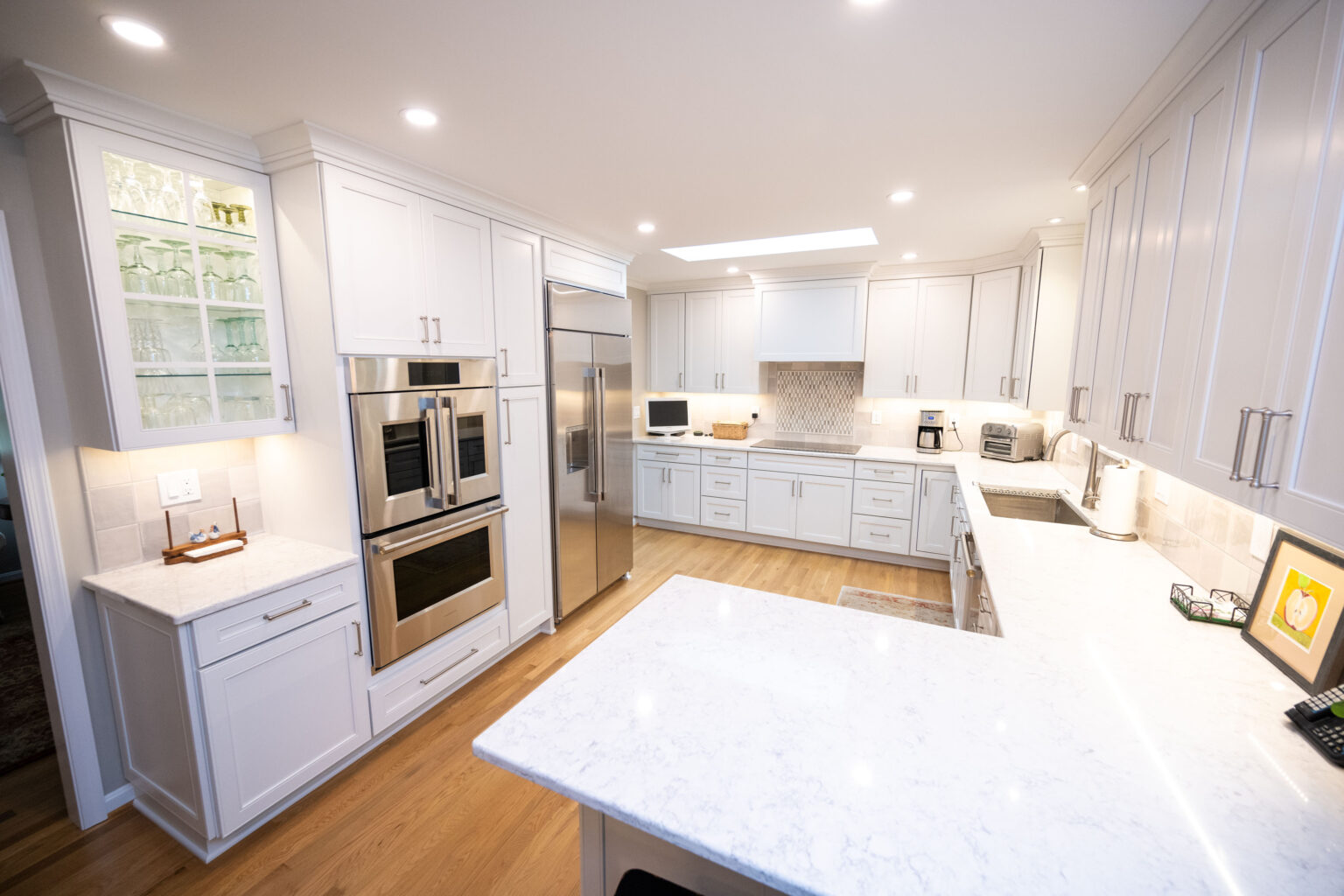 Image by Lesnick Photo A bright, modern kitchen with white cabinets, stainless steel appliances, and marble countertops, featuring a double oven, built-in microwave, and various small kitchen appliances.