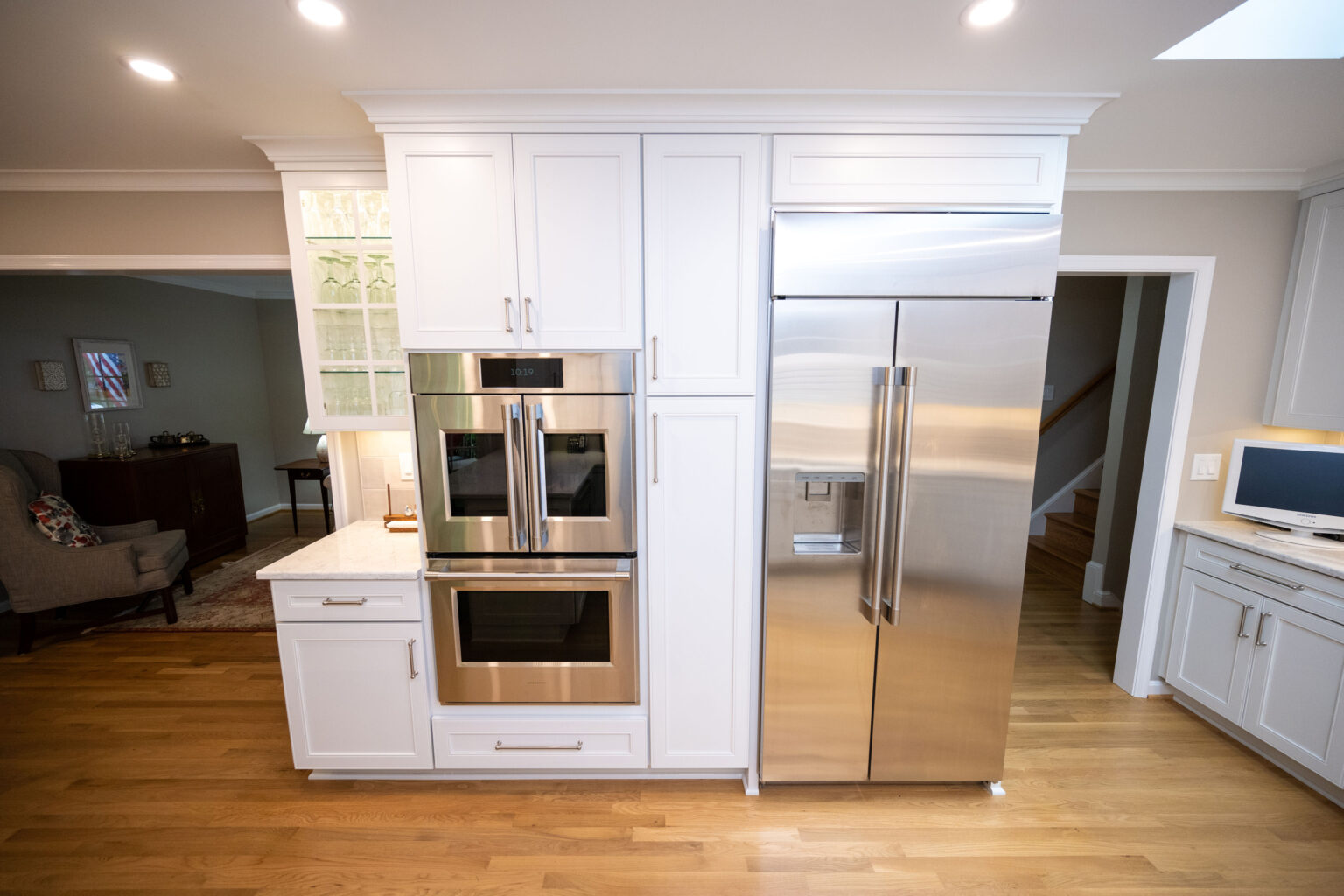 Image by Lesnick Photo A modern kitchen featuring white cabinets, double wall ovens, and a large stainless steel refrigerator on hardwood floors. A small desk area is visible to the right.