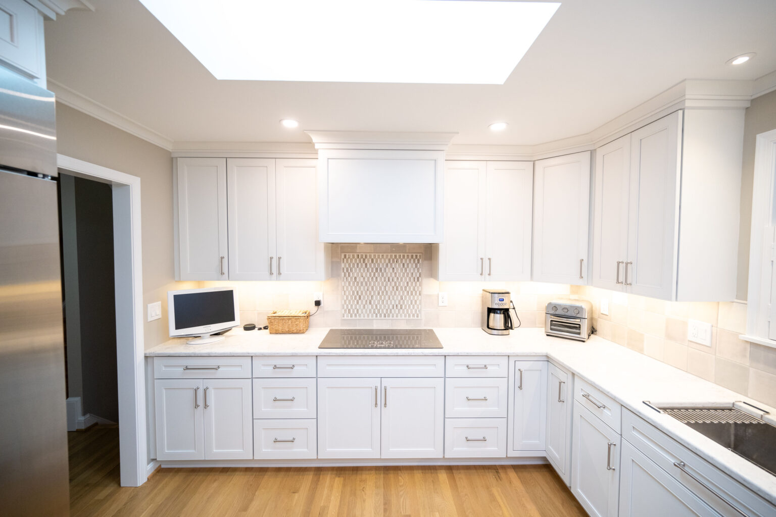 Image by Lesnick Photo A modern kitchen with white cabinets, wood flooring, a skylight, stainless steel appliances, and a basket on the counter near a small TV.