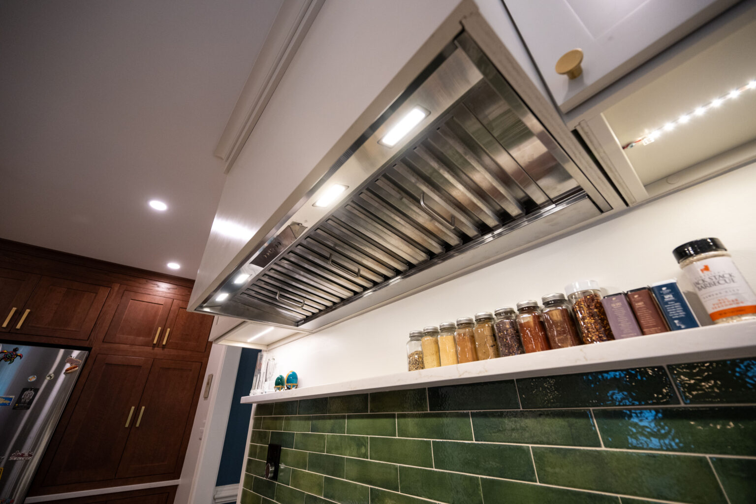 Image by Lesnick Photo Stainless steel kitchen range hood with lights above a tile backsplash, spice jars, and a cabinet with wooden doors and a refrigerator in the background.