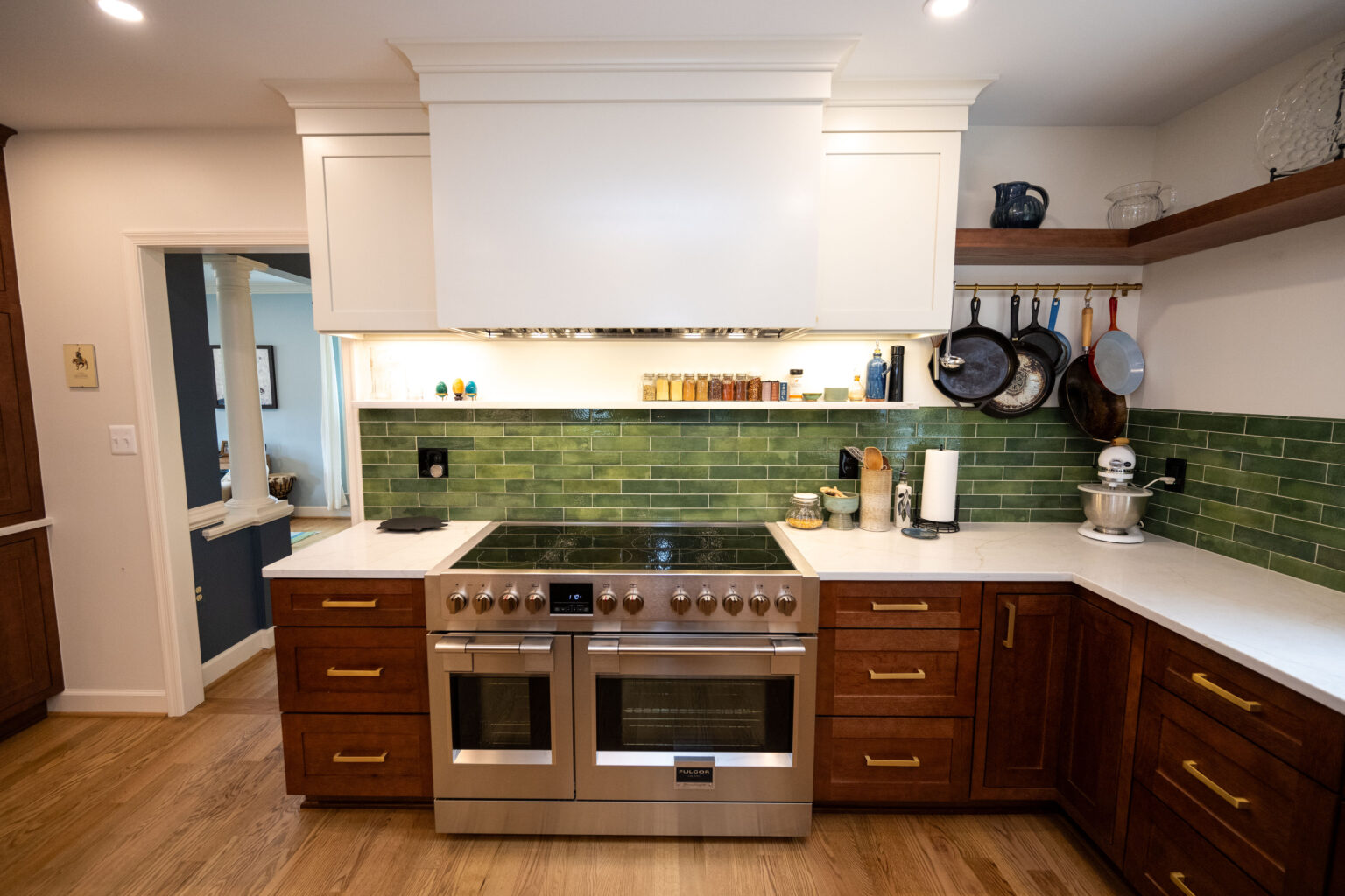 Image by Lesnick Photo Modern kitchen with a stainless steel double oven, green tile backsplash, and wooden cabinets. Cooking utensils and jars are on the shelves. Wooden flooring is visible.