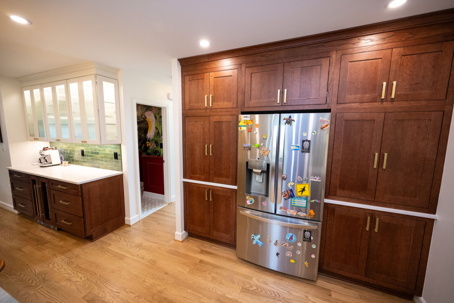 Image by Lesnick Photo Modern kitchen with wooden cabinets and a stainless steel refrigerator decorated with magnets. A small alcove with green backsplash and a visible kitchen appliance is in the background.