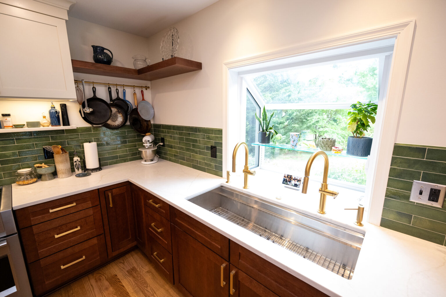Image by Lesnick Photo A modern kitchen with green tile backsplash, double sink under a window, wooden cabinets, and pots hanging above the countertop. Bright natural light illuminates the space.
