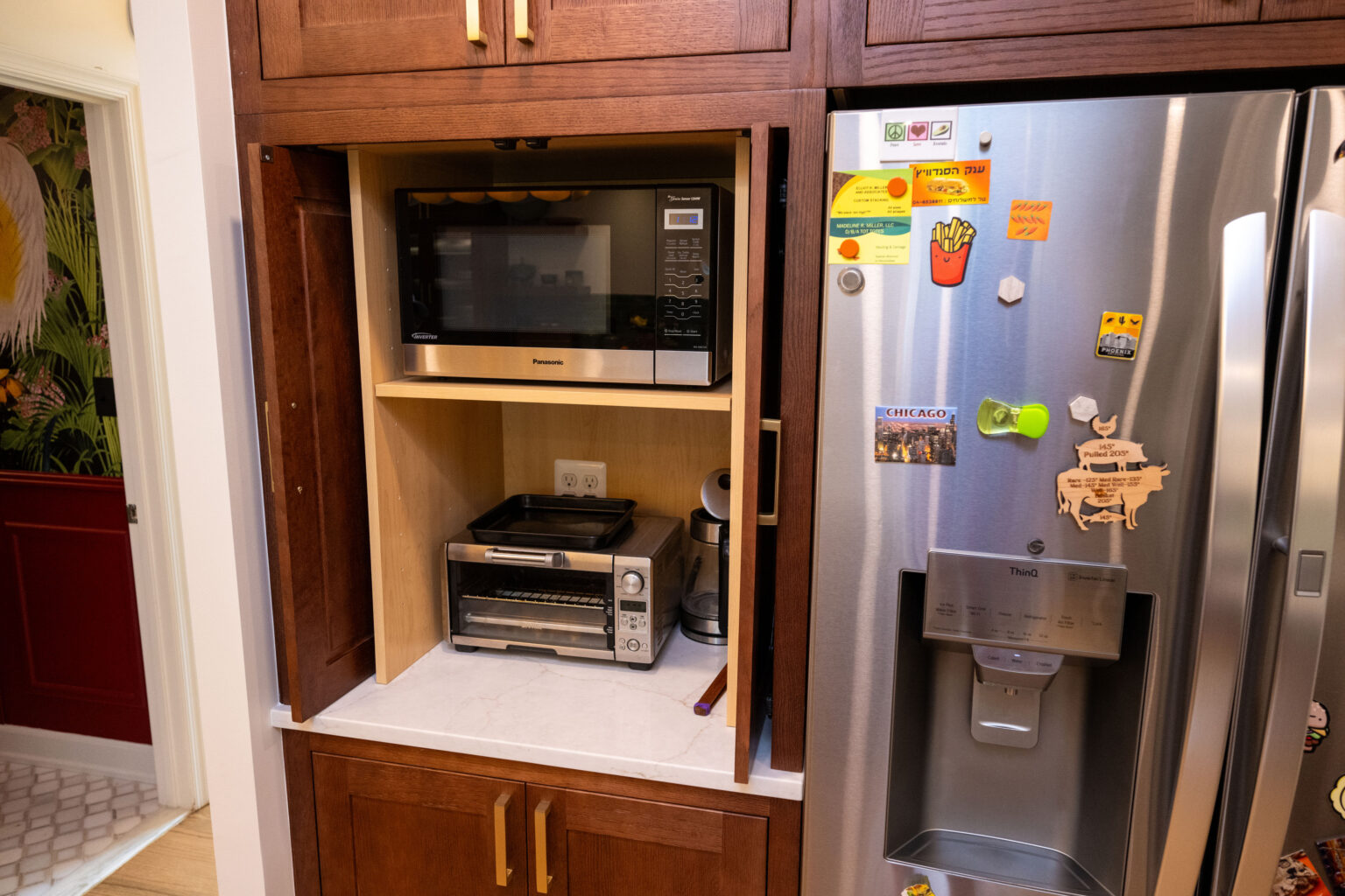 Image by Lesnick Photo Kitchen appliances, including a microwave and toaster oven, are neatly placed in a wooden cabinet next to a stainless steel refrigerator decorated with colorful magnets.
