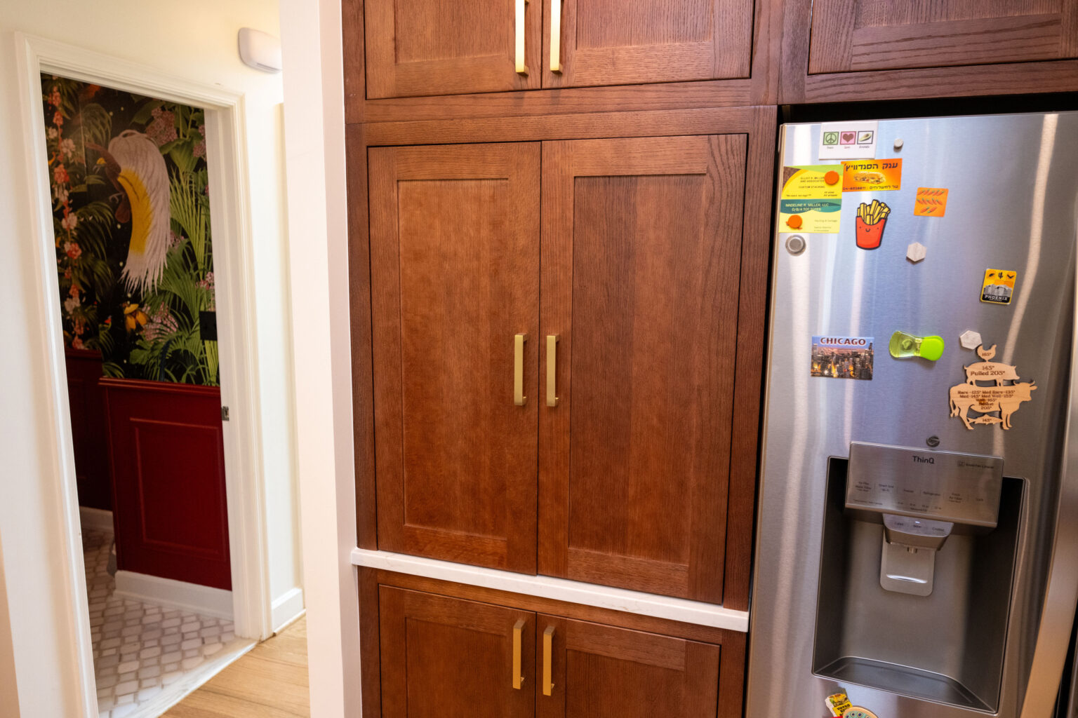 Image by Lesnick Photo Kitchen interior with wooden cabinets and a stainless steel refrigerator covered in magnets. A door on the left opens to a room with decorative wallpaper.