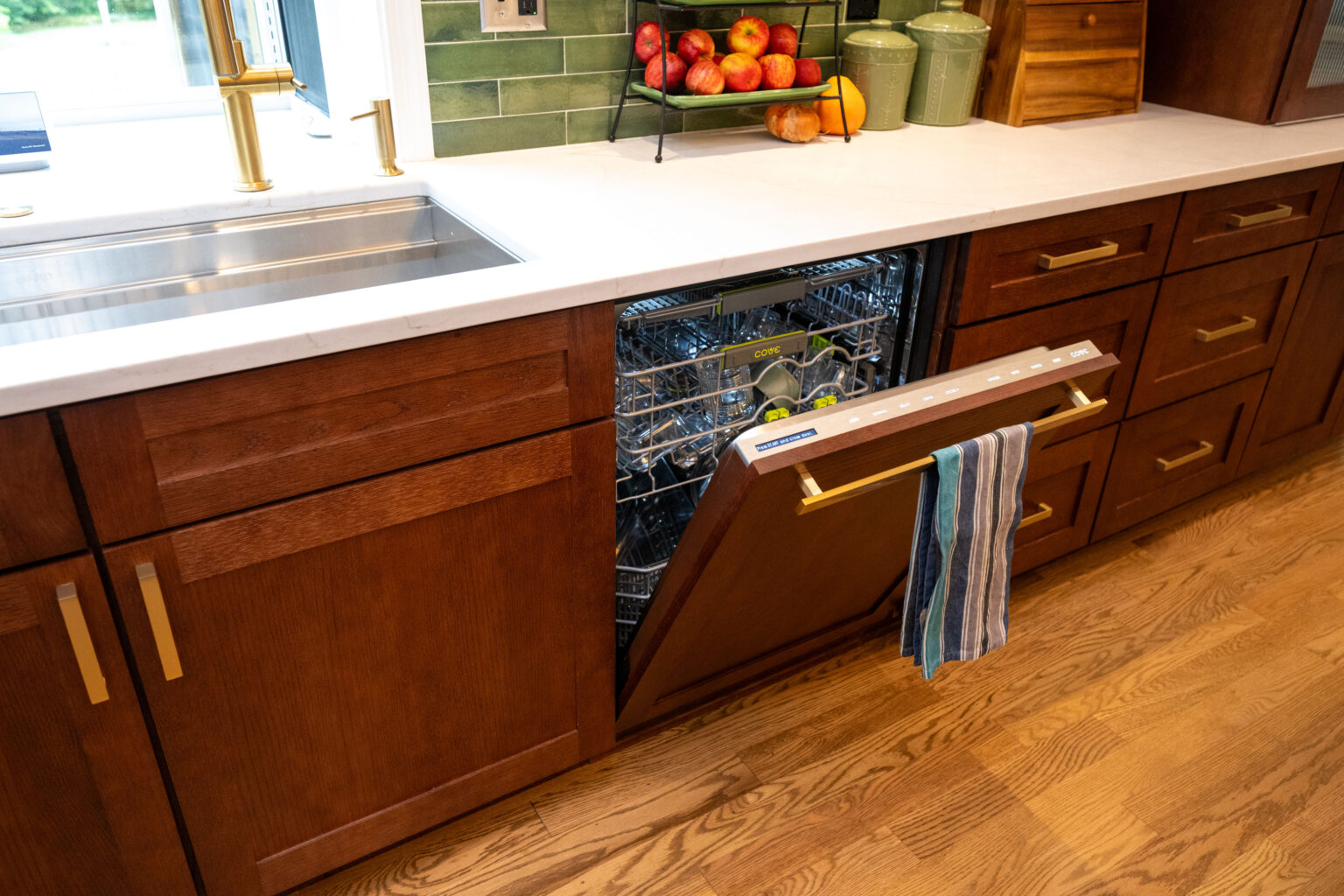 Image by Lesnick Photo A kitchen with wooden cabinets features an open dishwasher, a striped towel hanging on its door, and a countertop with a fruit tray, green jars, and a gold faucet by the sink.