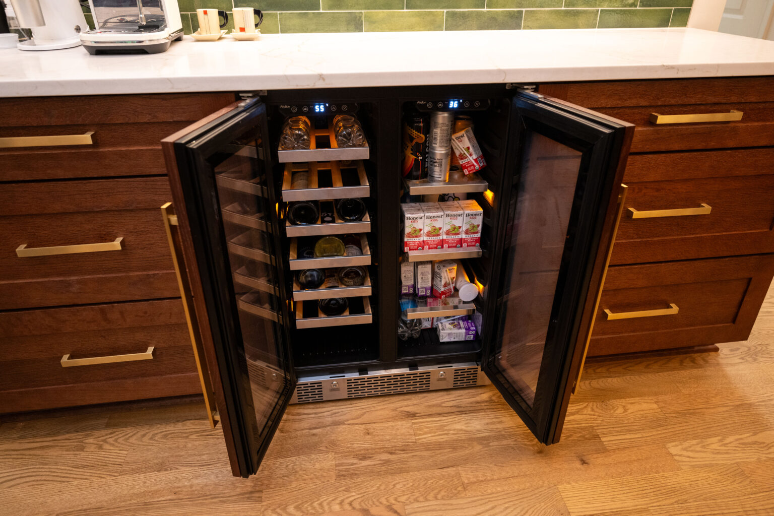 Image by Lesnick Photo A dual-zone wine fridge is open, displaying bottles of wine on the left and various boxed drinks and snacks on the right, set within a wooden kitchen cabinet.