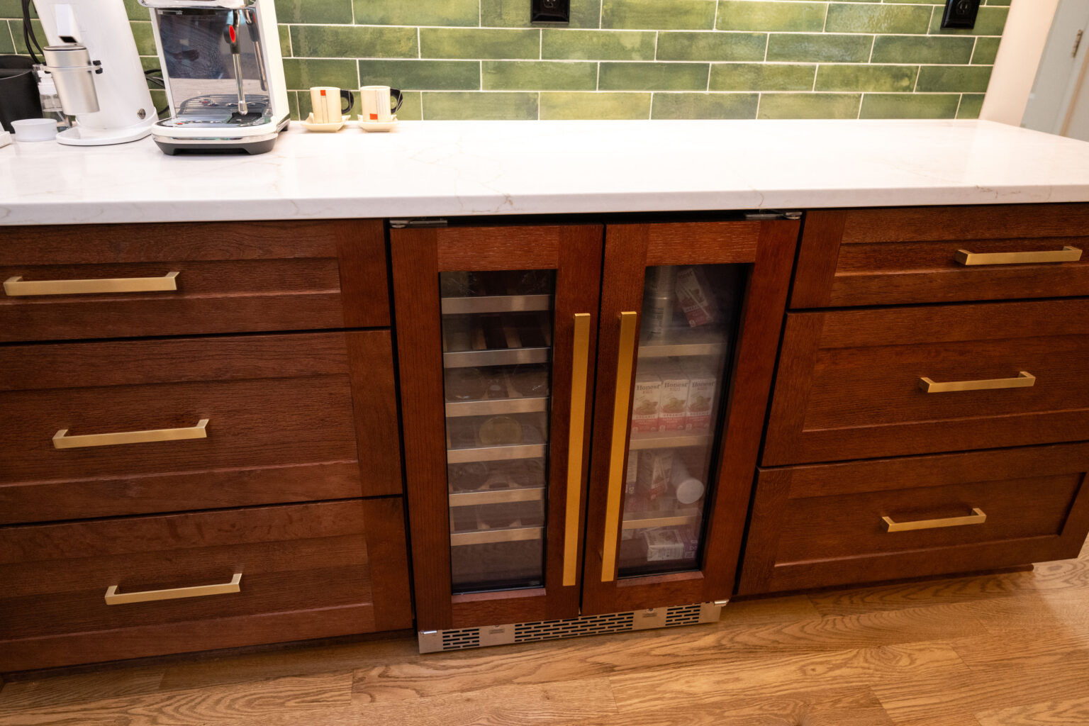 Image by Lesnick Photo Wooden kitchen cabinets with gold handles, a marble countertop, and a glass-door mini fridge beneath. A coffee maker sits on the counter against a green tiled backsplash.