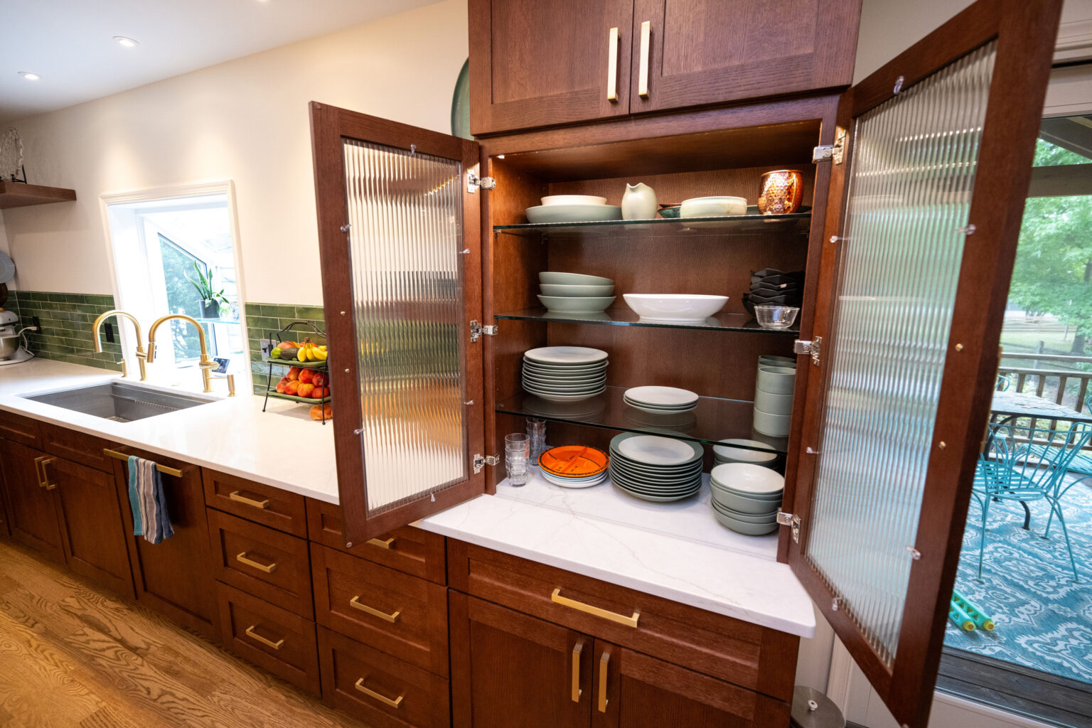 Image by Lesnick Photo Open kitchen cabinet with glass doors displaying neatly stacked dishes and bowls. Wooden cabinets, marble countertop, and a view of a dining area with greenery outside.