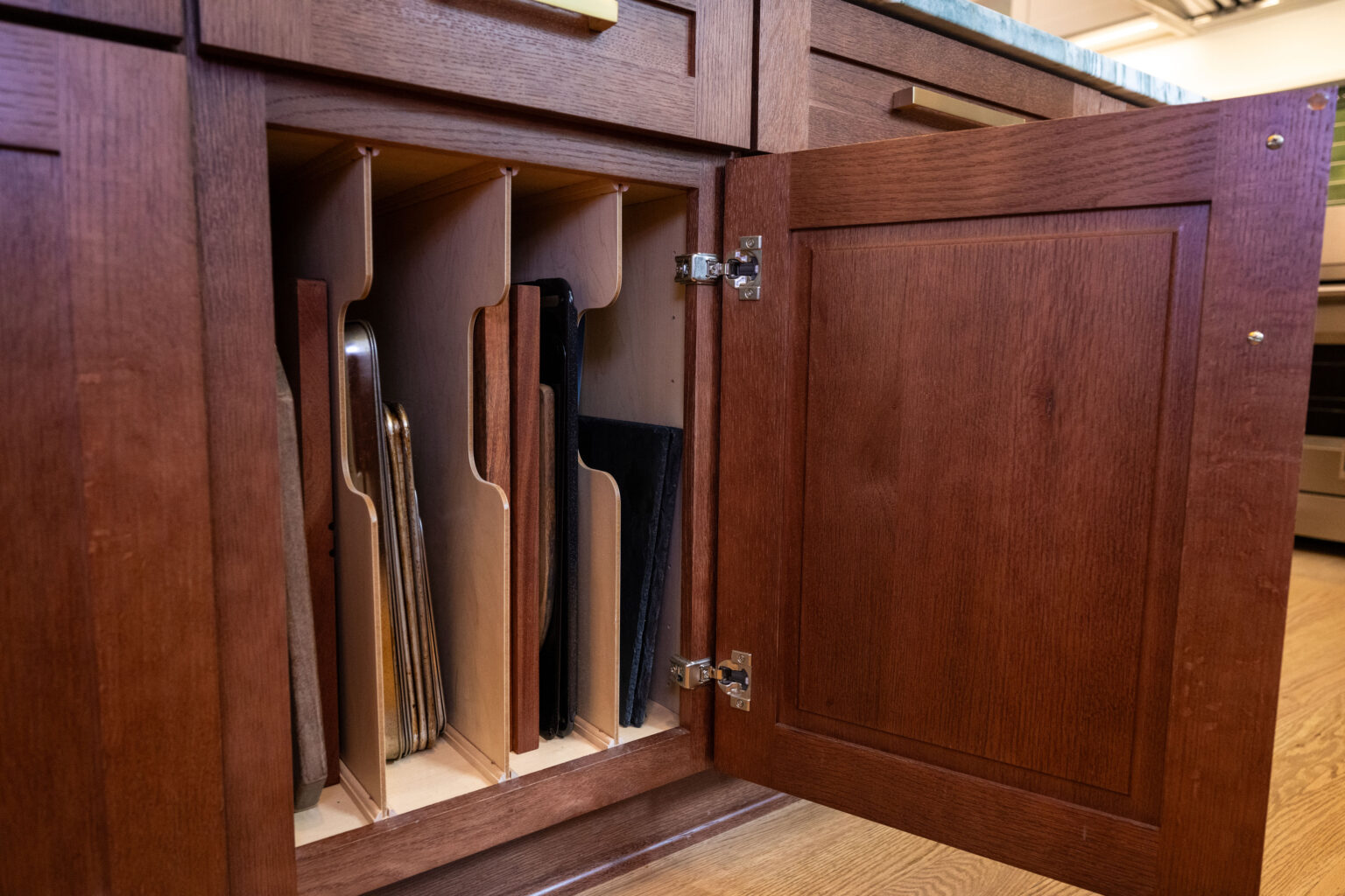 Image by Lesnick Photo A wooden kitchen cabinet with an open door reveals vertically stored cutting boards and baking sheets.
