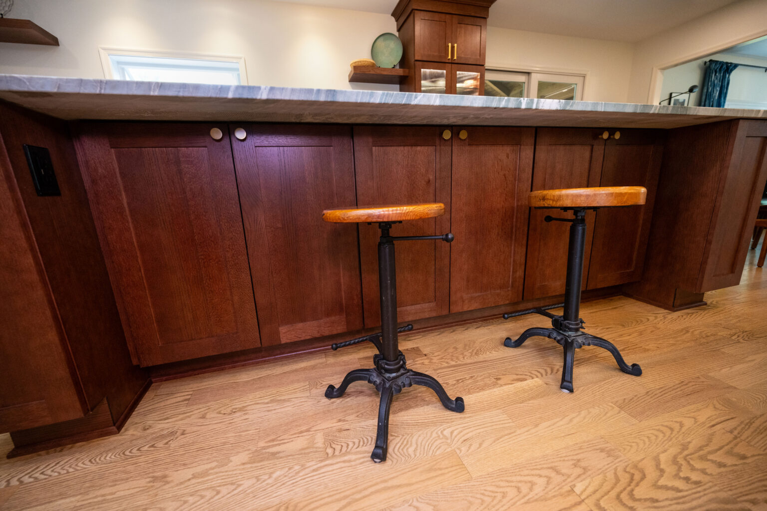Image by Lesnick Photo Two wooden stools with metal bases are positioned in front of a kitchen island with dark wood cabinets and a granite countertop.