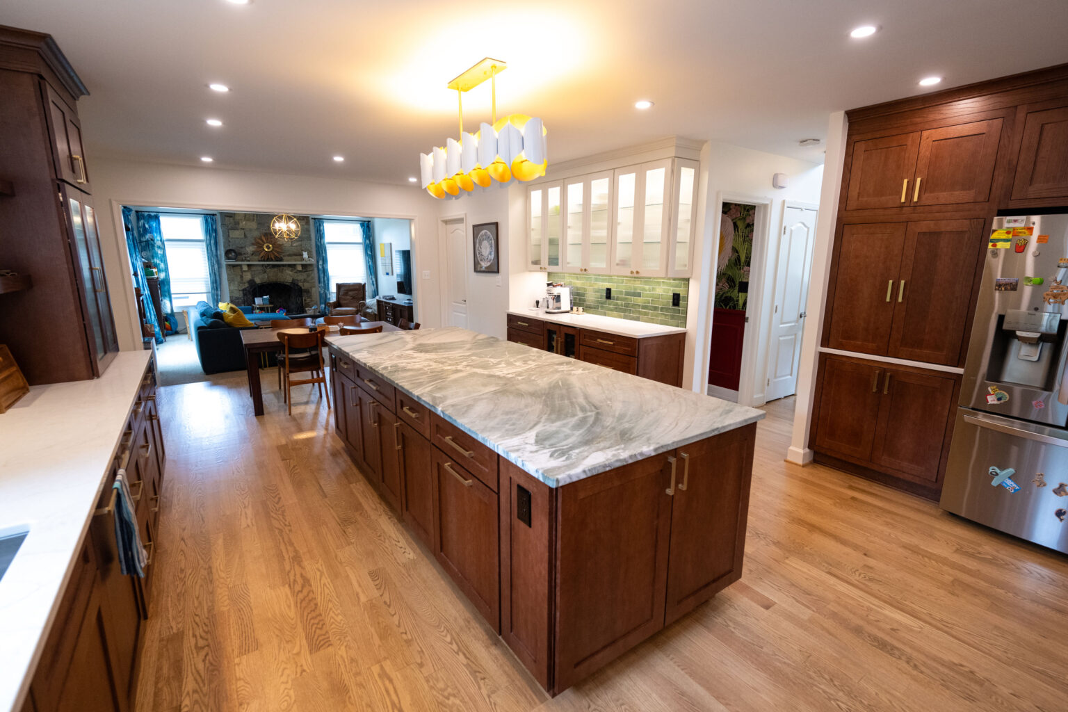 Image by Lesnick Photo Modern kitchen with a large marble island, wooden cabinets, and stainless steel fridge. A dining area with a table and chairs is visible in the background.