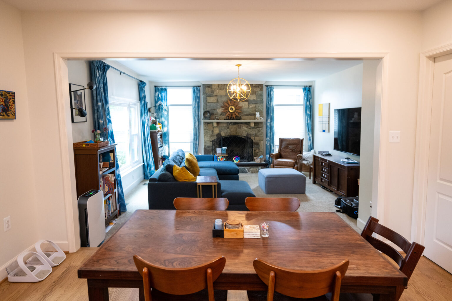 Image by Lesnick Photo A cozy living room with a dining table and four chairs in the foreground. A stone fireplace, sofas, a TV, and a window are visible in the background. Brightly lit with a warm ambiance.