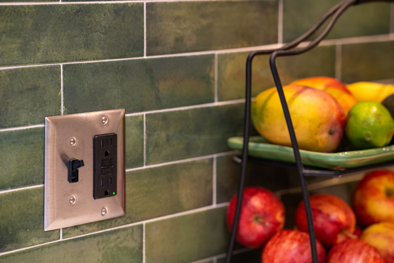 Image by Lesnick Photo Close-up of a kitchen wall with green tiles, featuring a stainless-steel electrical outlet. A fruit stand with apples, limes, and mangoes is partially visible on the right.